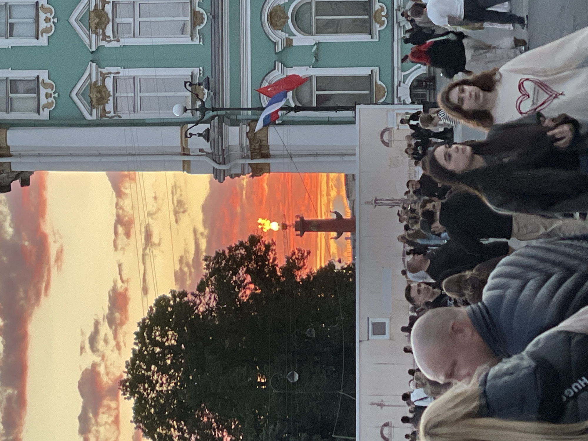 In an outdoor urban setting under a vibrant sunset, a crowd of people mill about in front of a large, ornate teal building with white architectural details and gold accents. A tall red column with a lit flame at its apex stands in the middle ground, silhouetted against the fiery orange and pink sky. A Russian flag and a red flag fly from a lamppost attached to the teal building. Several trees are visible, with one large, dark green tree to the left of the frame partially obscuring the sunset. The people in the foreground are diverse, with some facing away from the camera and others looking forward. Their clothing suggests a casual urban environment. The scene is illuminated by the warm glow of the setting sun and the flame atop the column.