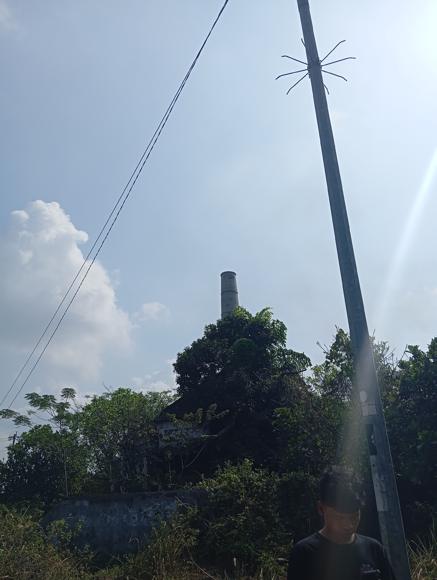 The image depicts an outdoor scene with a clear sky and sunlight. In the foreground, a young male wearing a black t-shirt is looking down at a phone, standing next to a utility pole. The utility pole has a star-shaped antenna-like structure attached to it. In the mid-ground, a tall, cylindrical smokestack rises above dense green foliage. The background consists of a blue sky with scattered white clouds. The environment appears to be a rural or semi-rural area, possibly on the outskirts of Sokaraja, Indonesia. The vegetation suggests a tropical climate.