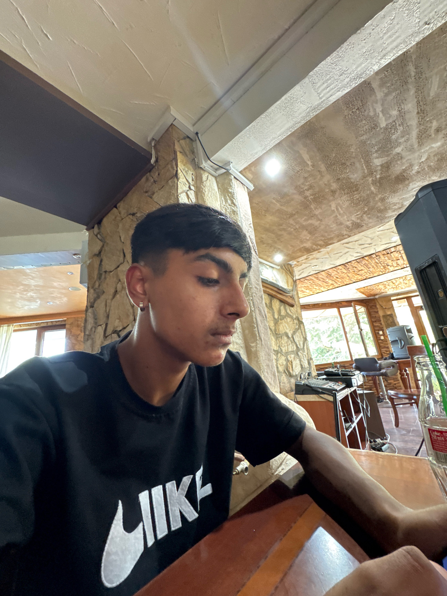 The image shows a young man with short dark hair, wearing a black Nike t-shirt, sitting at a wooden table. He is wearing a silver hoop earring in his left ear. His right arm rests on the table. The background suggests an indoor setting with exposed stone walls and a textured ceiling. Large windows are visible, revealing greenery outside. Several objects are present in the background, including what appears to be audio equipment, chairs, and a bottle with a green straw. The lighting is mixed, with natural light coming from the windows and artificial lights from the ceiling. The overall ambiance suggests a casual, possibly entertainment-oriented venue. The location is identified as Sredets, Bulgaria.
