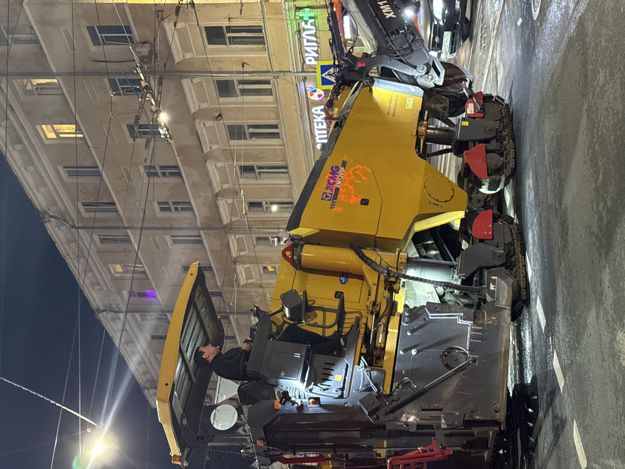 The image shows a road milling machine parked on a street in Nizhny Novgorod, Russia at night. A man is sitting in the driver's seat. The machine is yellow and gray, with the logo "XCMG" and the model number "XM1205F" visible on its side. The Cyrillic word "АПТЕКА" (pharmacy) and "РИГЛА" (Rigla, a pharmacy chain) are illuminated in neon lights on a building in the background. Overhead power lines are visible above the street. The road is wet and marked with white lane lines. Other cars are visible on the road.