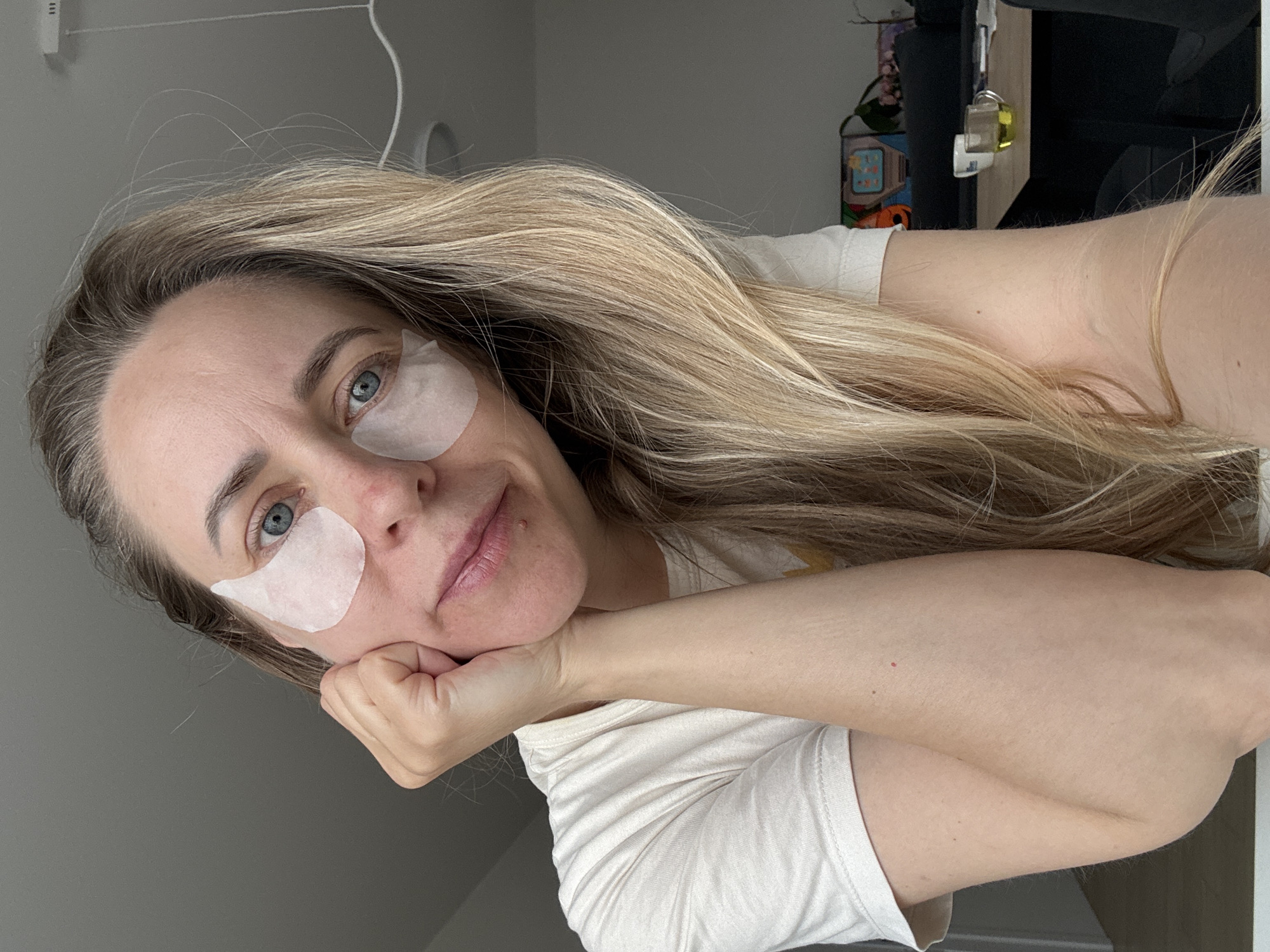 A fair-skinned woman with long blonde hair poses for a selfie. She is wearing white under-eye patches. She rests her chin on her hand. She is wearing a white t-shirt. The background shows a wall and partial view of furniture. The location is not discernible from the image.
