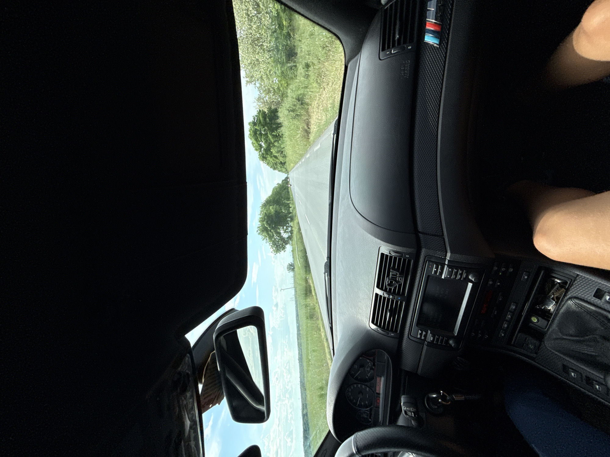 The view from inside a car shows a road stretching ahead with trees lining either side. The car's dashboard, vents, rearview mirror, and steering wheel are visible. A person's leg is visible on the right side of the frame. The location, Karavelovo, Bulgaria, cannot be confirmed based on the image alone.