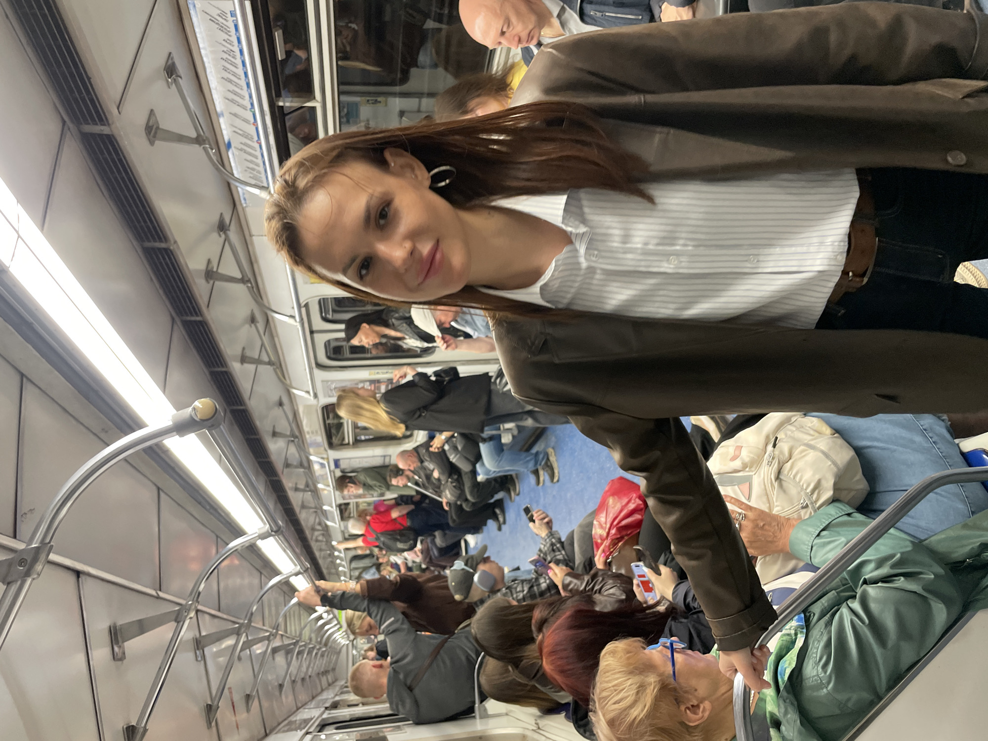 The image shows the interior of a subway car in an unknown city. The car is crowded with passengers of various ages, genders, and ethnicities. Some are sitting, while others are standing and holding onto the handrails. Many are looking at their phones.
The woman in the foreground is looking directly at the camera with a slight smile. She has brown hair and is wearing a light-striped shirt under a brown jacket.