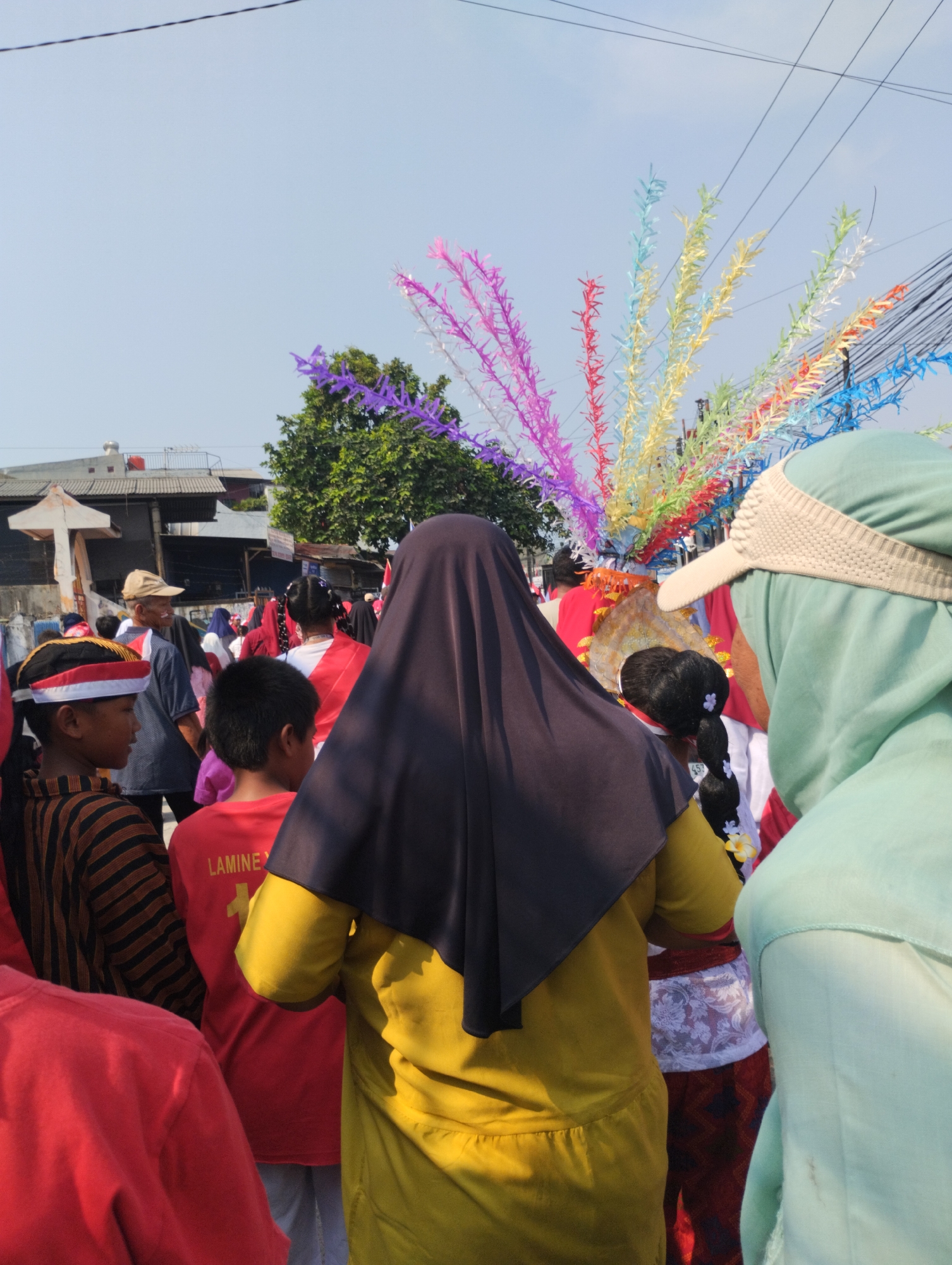 In a scene typical of a local festival or celebration in Bekasi, Indonesia, a group of people are gathered outdoors under a bright, clear sky. The focal point is a colorful, fan-like decoration made of bright, slender strips of paper in various colors including pink, purple, light blue, yellow, and white, which rises high above the crowd.
In the foreground, from left to right, is the back of a person wearing a bright red shirt. To their right, a young boy with a striped shirt and a red and white head covering is looking to his left. Next to him, a boy in a red t-shirt, which has text "LAMINE Y" visible on the back, is looking forward. A person in a dark head covering and a bright yellow garment is positioned centrally, their back to the camera. To their right, a person wearing a light green head covering and a beige visor is seen from the side. Further in the background, several other individuals are visible, some wearing red and white outfits, and a person in a beige cap.
The environment appears to be a somewhat urban or suburban area, with buildings and structures visible in the background, including what looks like a white cross-like structure on the left. Overhead, power lines crisscross the sky. The overall atmosphere suggests a community event, possibly a parade or cultural gathering, in Bekasi.