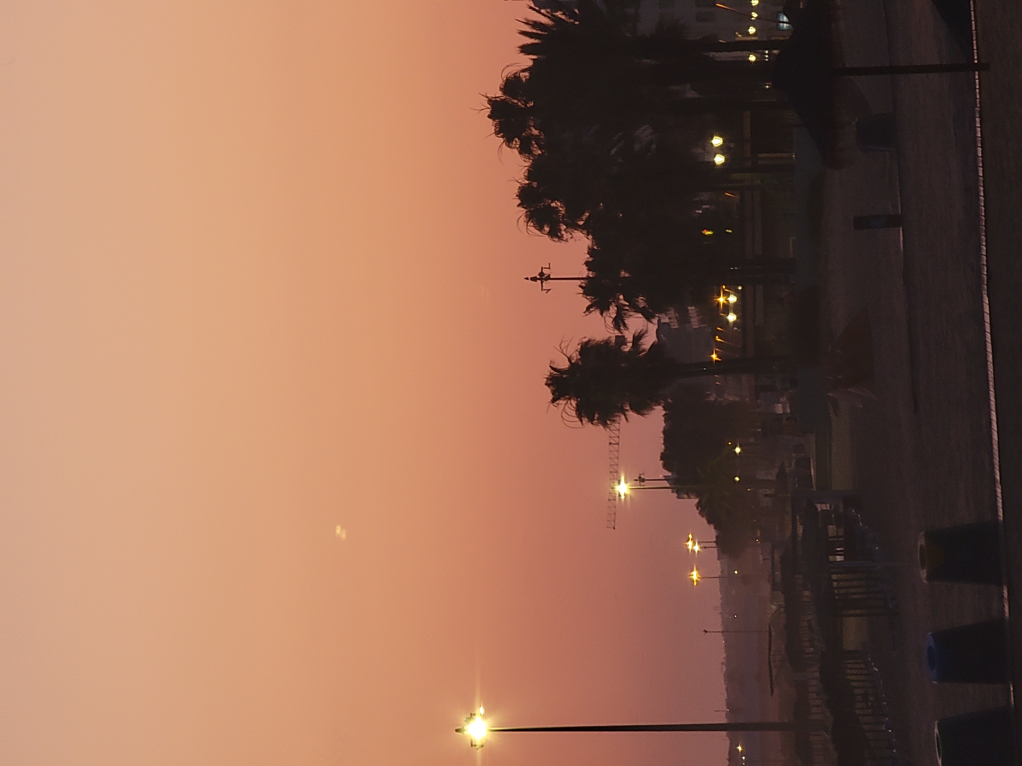 A vertical, low-angle shot captures a beach scene at dusk or dawn in Quarteira, Portugal. The sky is a soft gradient of orange and pink. In the foreground, three dark, conical trash receptacles are visible. Beyond them, a dark, sandy beach stretches out, dotted with shaded beach structures and umbrellas. Palm trees and buildings, silhouetted against the sky, line the background. Streetlights and other illuminated objects punctuate the scene, casting a warm glow. The overall impression is one of a tranquil, coastal environment preparing for or recovering from a busy day.