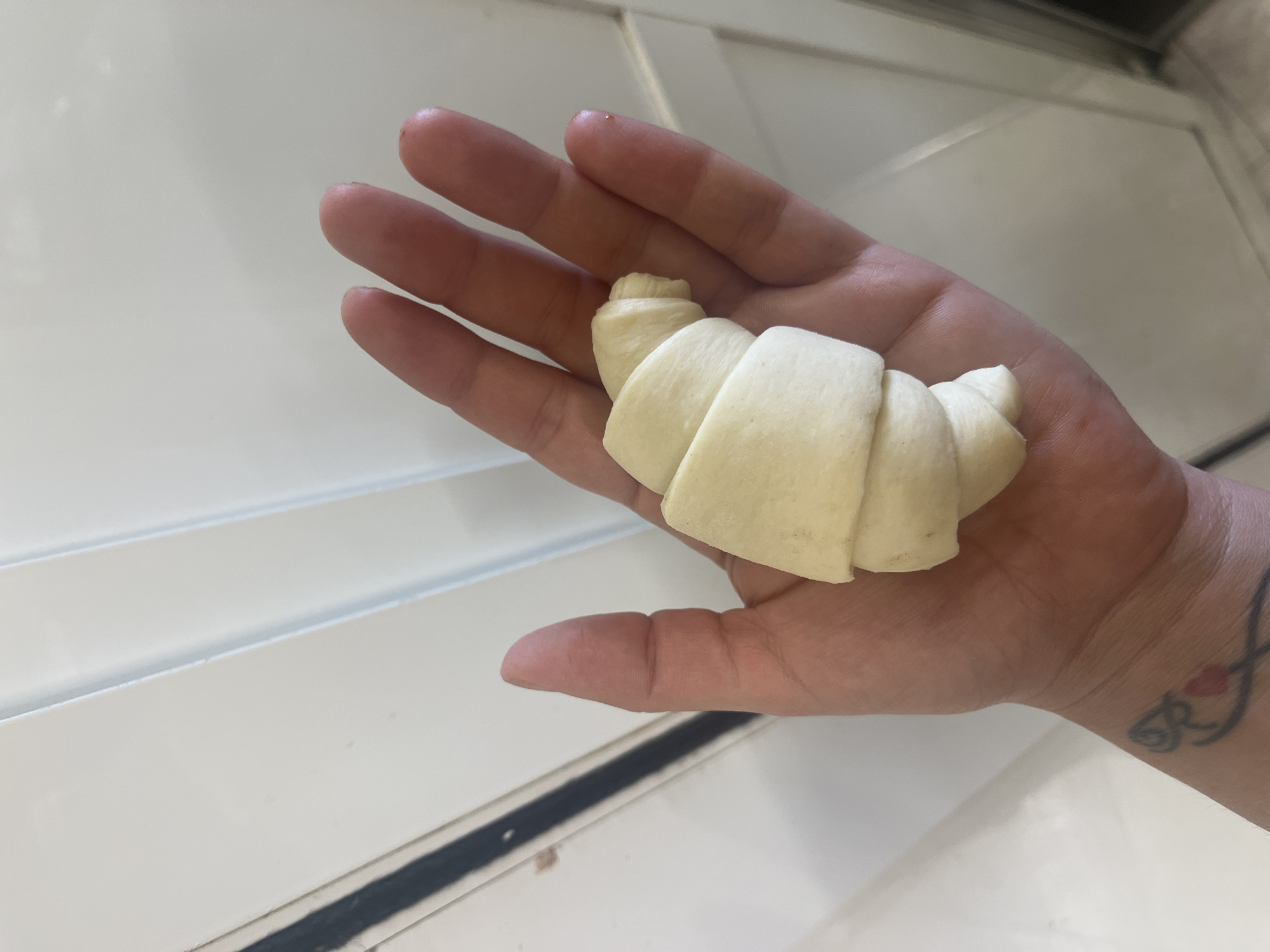 A human hand is holding a raw croissant. The hand is open, palm up, and the croissant is resting on it. The croissant is light yellow and appears to be made of dough. The hand has a tattoo on the wrist. The background is a white, tiled surface. The image is taken from a top-down perspective. There is no information about the location in Vallo della Lucania, Italy.