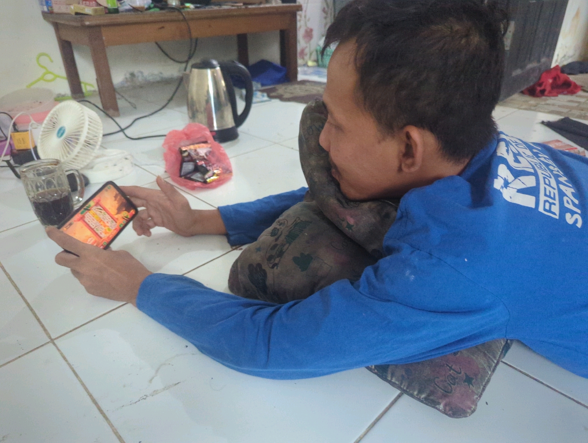 In Batu Belaman, Indonesia, a man lies on a tiled floor, propped up by a patterned cushion. He wears a blue long-sleeved shirt with white lettering on the back, partially obscured. His head rests on another cushion, and he holds a smartphone in his right hand, the screen displaying a game with colorful graphics. In the foreground, to the left of the man, a clear glass mug filled with a dark beverage sits on the floor, next to a small white fan. Behind the fan, a pink plastic bag contains some items, and further back, a stainless steel electric kettle is visible. A wooden table with objects on it is in the background, along with a wall and a floor with light-colored tiles. The overall scene suggests a moment of relaxation or leisure.