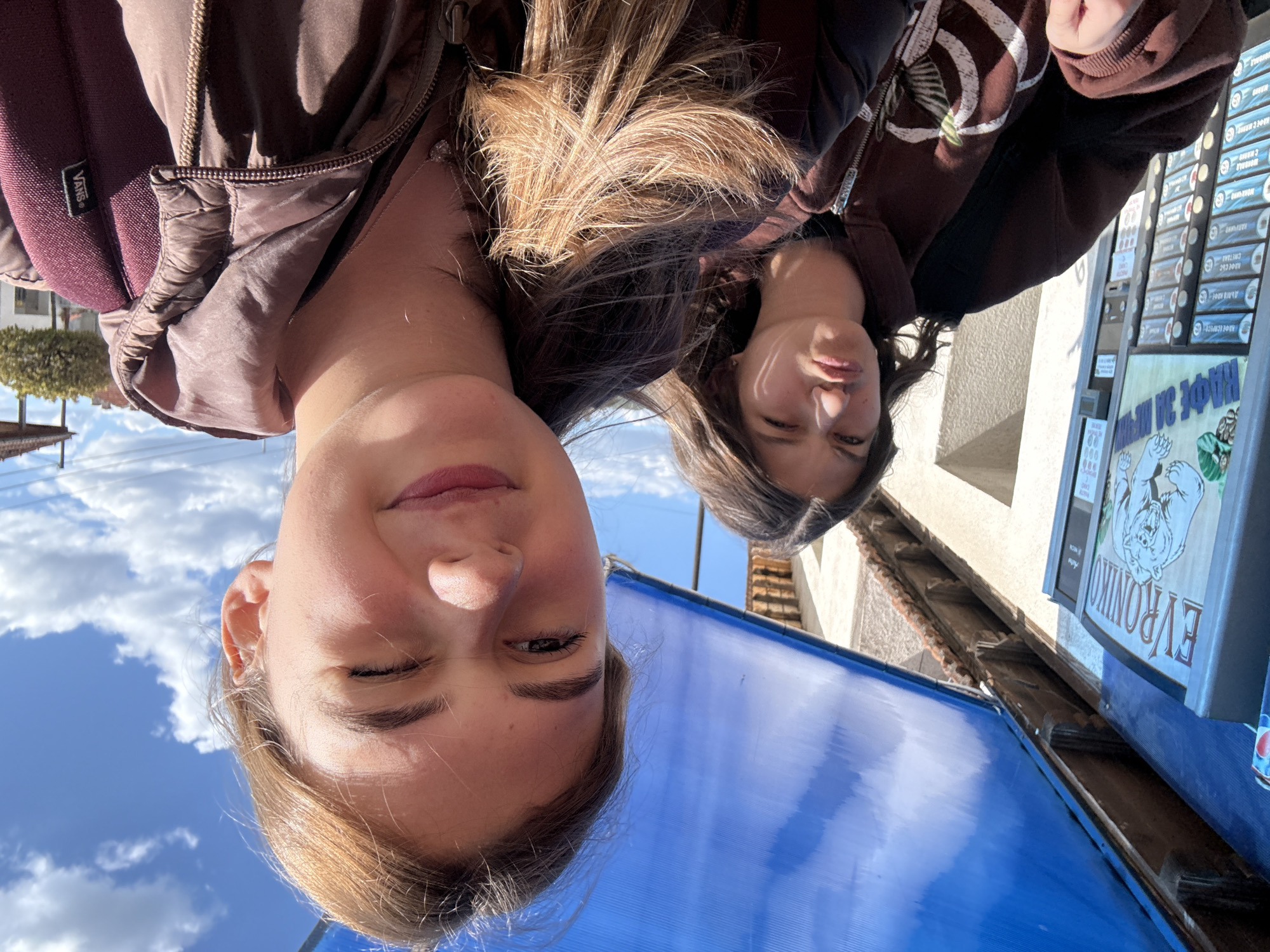 The image captures two young women outdoors, viewed from a low angle as they look up towards the camera. The woman in the foreground, positioned slightly to the left, has light brown hair, fair skin, and wears a brown puffer jacket over a dark red backpack with a "Vans" logo. She exhibits a subtle, closed-mouth smile. To her right, the second woman has darker hair and wears a brown hoodie with white patterns, displaying a more neutral or slightly pensive expression.
The background reveals a bright, clear blue sky dotted with white clouds, suggesting it is daytime, likely midday or early afternoon given the strong sunlight. Below the subjects, a prominent blue corrugated plastic awning extends across the lower portion of the frame. To the right, against a light-colored building wall, there's a vending machine or display stand featuring numerous items with Cyrillic text, hinting at various coffee or beverage options, with "КАФЕ" (Coffee) clearly visible on some labels. Adjacent to this, a larger sign displays a white bear logo, the Cyrillic text "EVRONIKО", and "КАФЕ ЗА ВЪНШНИ" (Coffee for Takeaway). A Pepsi can is also visible next to the sign. Further back, more buildings with tiled roofs and some trees can be seen.
The Cyrillic text on the signs and vending machine, along with the architectural style, are consistent with the specified location of Panagyurishte, Bulgaria. The women appear to be posing for a casual outdoor photograph.