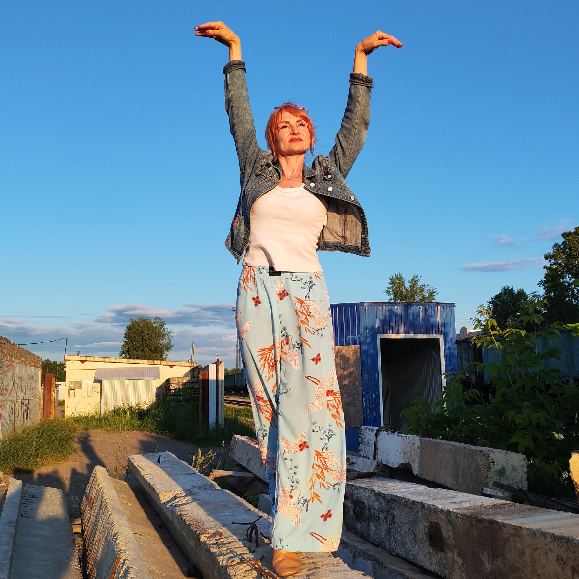 A woman with short, reddish-pink hair stands on a pile of concrete beams in an outdoor industrial or construction area, likely in Chaikovsky, Russia. She is wearing a denim jacket over a white t-shirt and loose-fitting, light blue pants with a pattern of cranes and orange accents. Her arms are raised above her head with her hands open, as if in a pose of celebration or surrender.
The scene is set against a clear, bright blue sky with a few wispy clouds. The sun appears to be low in the sky, casting long shadows and highlighting the textures of the concrete. In the background, there are various structures, including a blue industrial building with an open doorway, a long, light-colored building, and some vegetation. The overall impression is one of an informal, perhaps artistic, moment captured in a somewhat unkempt environment. The lighting suggests it is late afternoon or early evening. There is no visible text in the image.