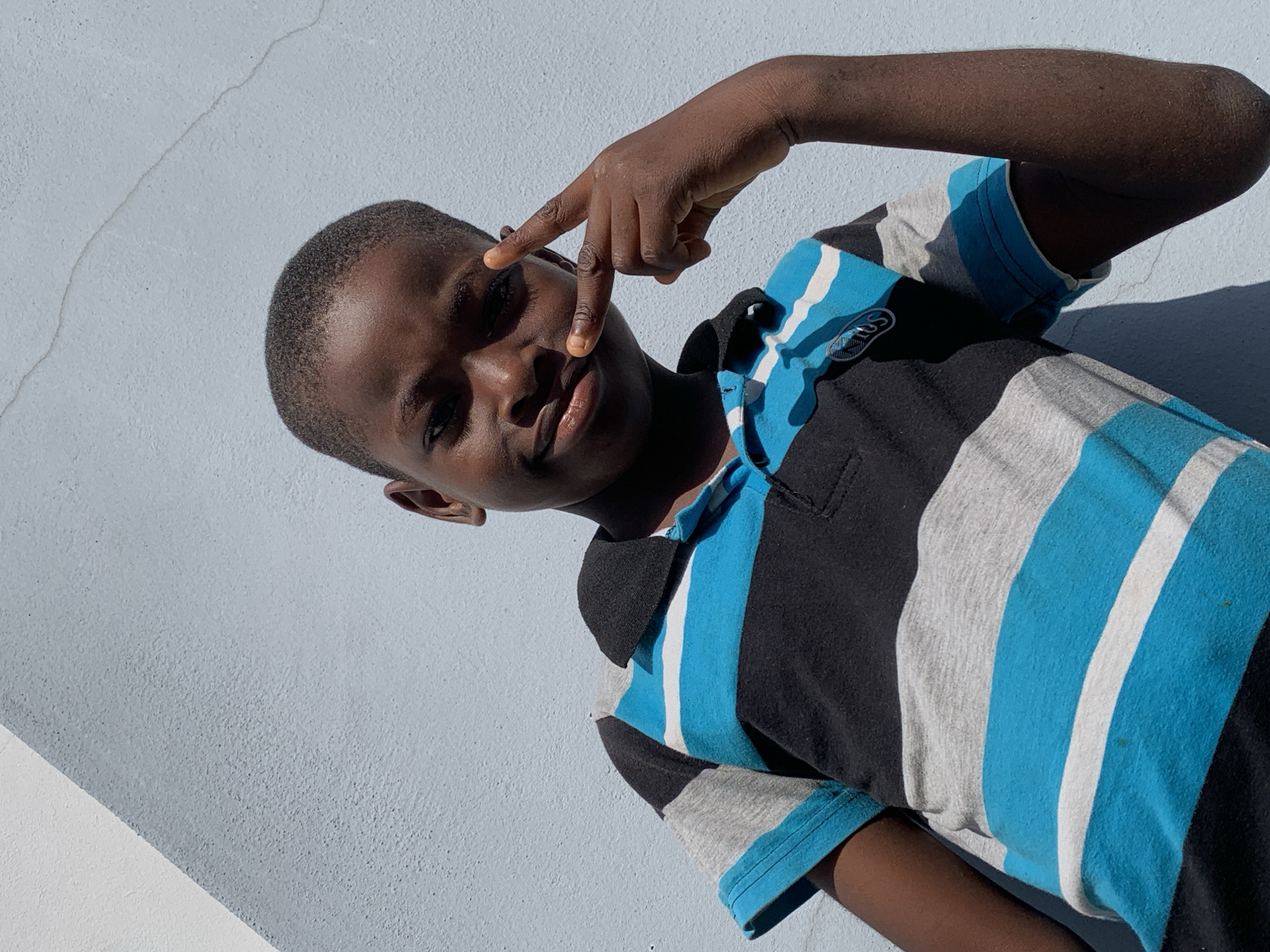 A close-up portrait shows a young boy with dark skin, positioned at an angle against a light blueish-grey textured wall. He is looking directly at the camera with a slight smile or smirk, while making a peace sign (V-sign) with his right hand, held near his left eye. The boy has short, dark hair and is wearing a short-sleeved polo shirt with distinct vertical stripes in black, gray, bright blue, and white.
Bright, direct sunlight illuminates the scene, casting strong, well-defined shadows on the textured wall behind him, indicating an outdoor setting during the day. A faint, thin crack is visible on the wall's surface in the upper left portion of the image. On one of the blue stripes of his shirt, near the left shoulder, there is a small white oval-shaped logo with unidentifiable text. The boy's confident and playful expression is notable. The scene is set in Ibasa, Nigeria.