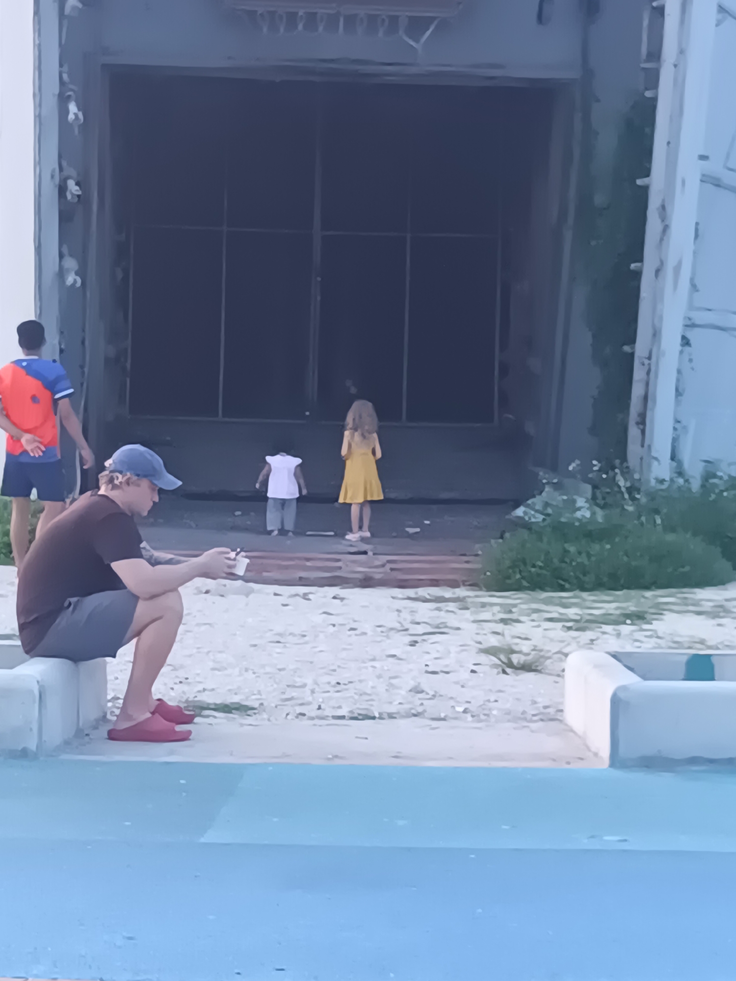 In a scene possibly located in Ko Phangan, Thailand, a man wearing a blue baseball cap and a dark t-shirt is sitting on a concrete ledge, holding a small white cup and looking down at his phone. He is wearing grey shorts and red slides. To his left, a person wearing a blue shirt with orange and red accents and dark shorts stands with their hands behind their back. In the background, a large, dark, industrial-style doorway is visible. Two children are standing near the doorway: one child in a white dress and grey pants, and another child in a yellow dress. The ground in front of the doorway is covered in small stones and sparse vegetation. The scene appears to be outdoors, with a bright, overcast sky.