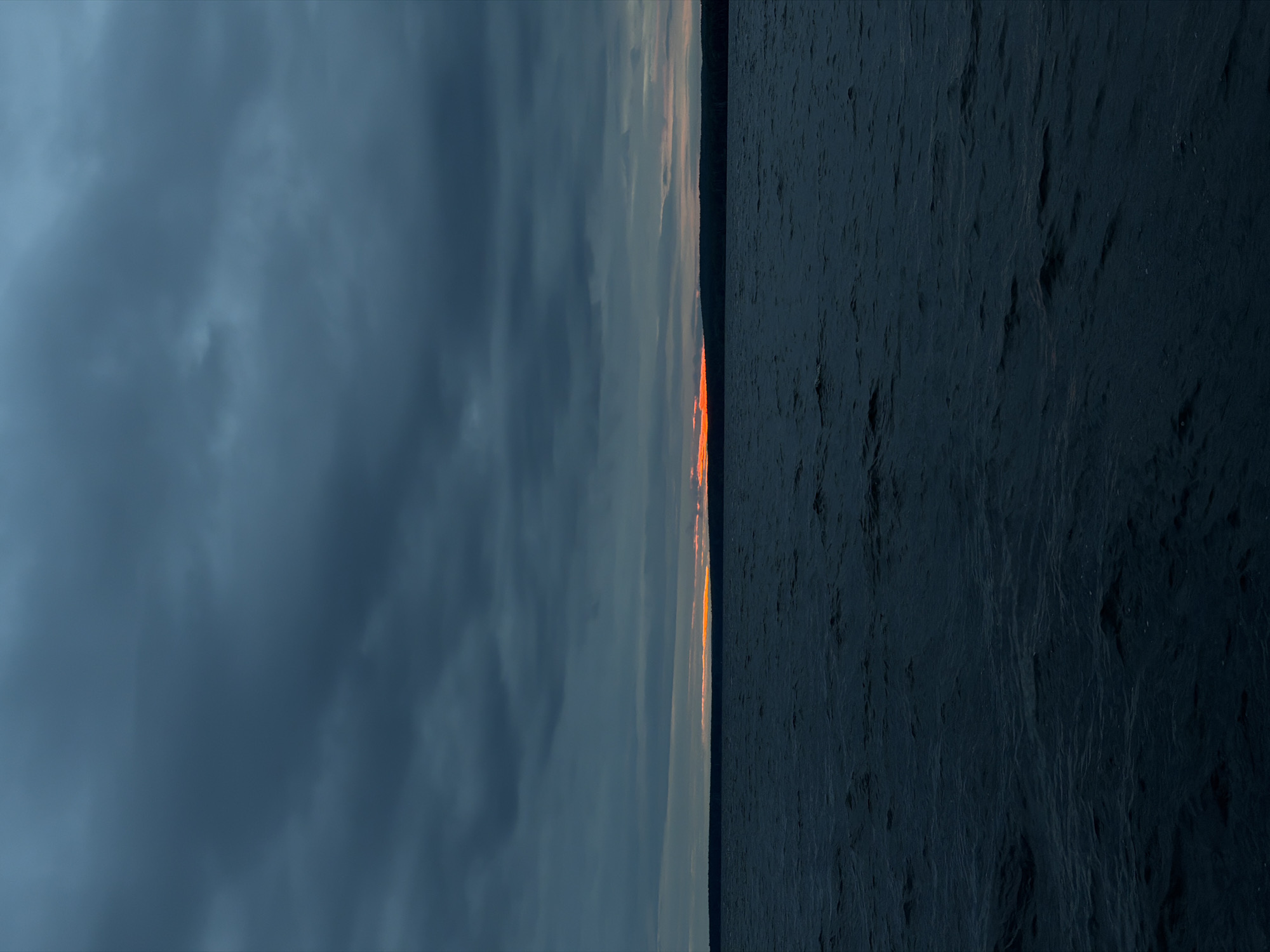 This image depicts a vast natural landscape in Chaikovsky, Russia, likely observed from a body of water, possibly the Votkinsk Reservoir. The upper two-thirds of the vertical frame are dominated by a dense, dark grey-blue cloud cover, indicating an overcast sky. A narrow, intense horizontal band of orange and red light is visible on the distant horizon, signifying either sunrise or sunset. This light band is partially obscured by a dark, silhouetted strip of land, presumably a forested shoreline. The lower third of the frame comprises dark, choppy water with visible ripples and small waves, reflecting the muted tones of the sky. No individuals, vessels, or man-made structures are present within the scene. The overall lighting conditions suggest twilight, creating a somber and expansive atmosphere.