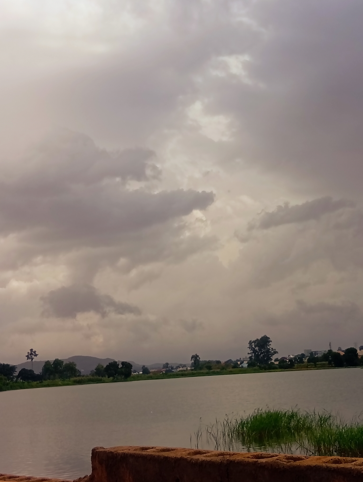 The image depicts a body of water, likely a lake or river, under a heavily overcast sky. The water is calm, reflecting the muted tones of the clouds. Along the far bank, a line of trees and vegetation is visible, with some scattered buildings and distant hills forming the horizon. In the foreground, the edge of a textured, reddish-brown structure, possibly a wall or embankment, is visible. The overall scene conveys a somber or tranquil atmosphere, typical of a cloudy day in a natural or semi-urban setting. The location is Udu, Nigeria.