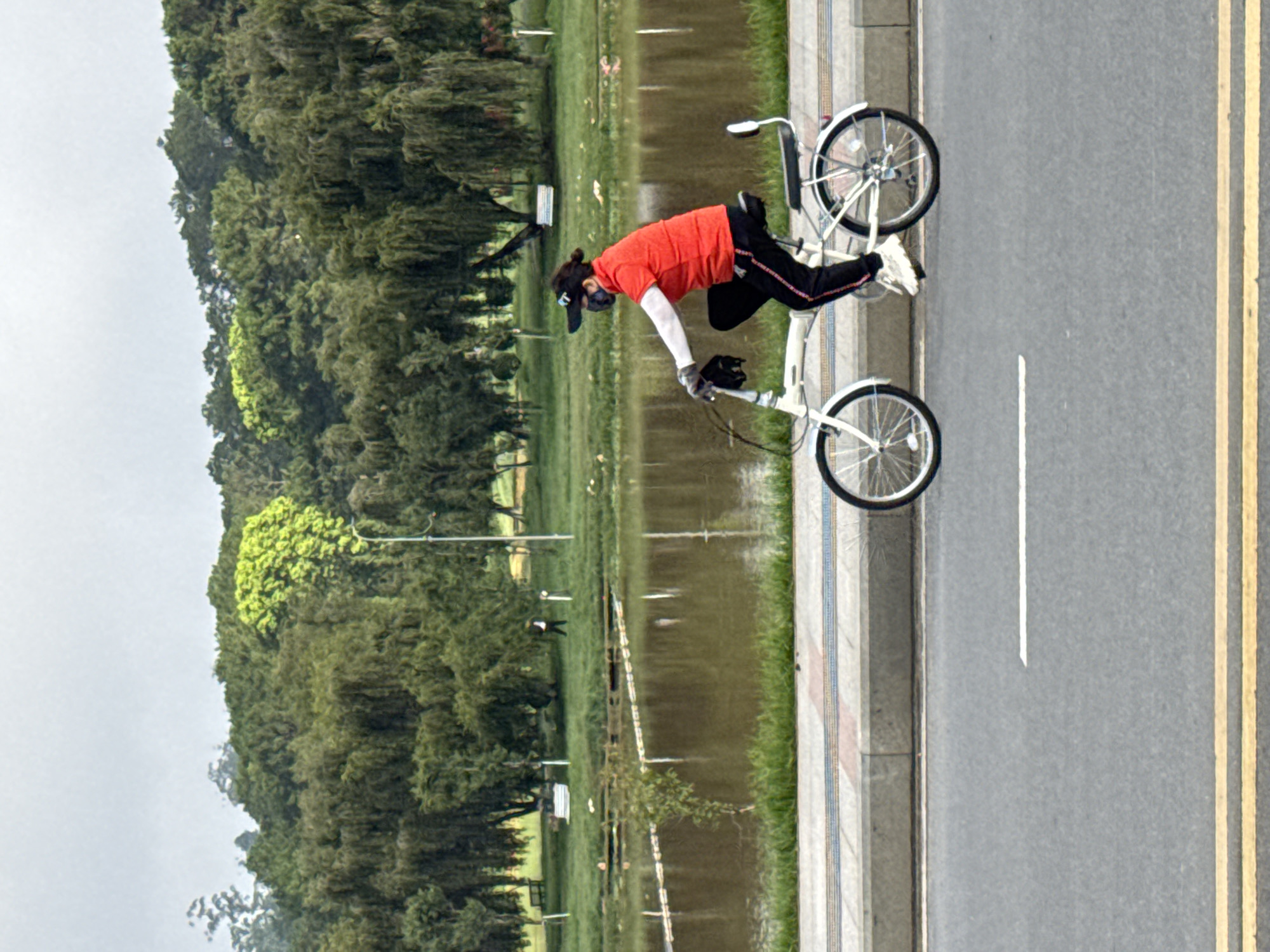 A person wearing a red vest, black pants, a black face mask, and a black cap rides a white folding bicycle along a paved path. The path is bordered by a sidewalk with a decorative stripe and a body of water with trees and a grassy area in the background. The scene appears to be a park or recreational area. This is likely Xuan Huong Lake in Da Lat, Vietnam, a popular spot for cycling and outdoor activities.