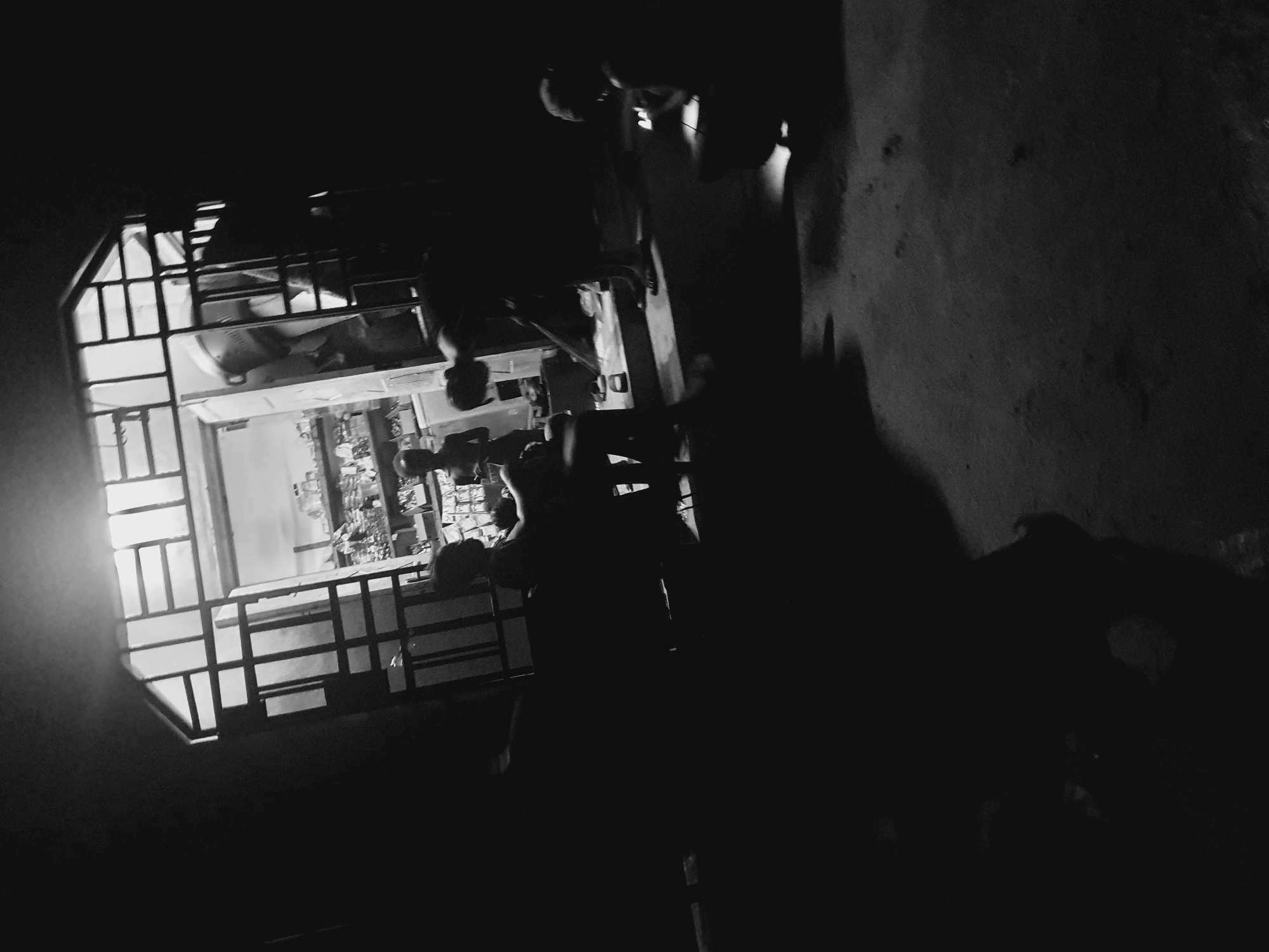 A black and white, low-angle, rotated view of a scene in Lokoja, Nigeria. The interior of a brightly lit room is visible through a window with a security grate. Outside the window, the scene is very dark. The silhouette of a person stands in the foreground. The wall to the right of the person is textured. It is night.