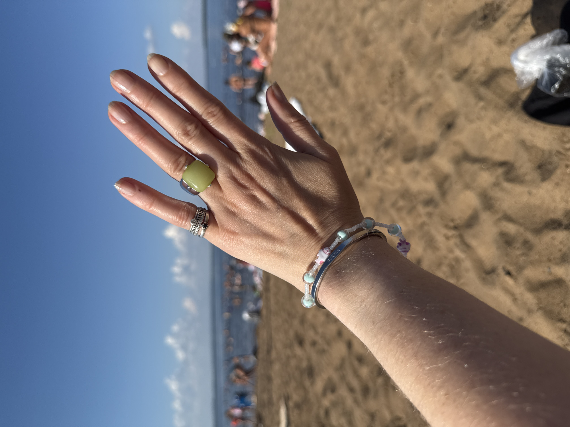 The image shows a person's arm and hand in the foreground, adorned with rings and bracelets. The person is on a sandy beach with a body of water and many people in the background. The sky is blue with some clouds. The scene appears to be a public beach during the daytime. This location is likely a beach on the Kama River in Chaikovsky, Russia, given the context provided.