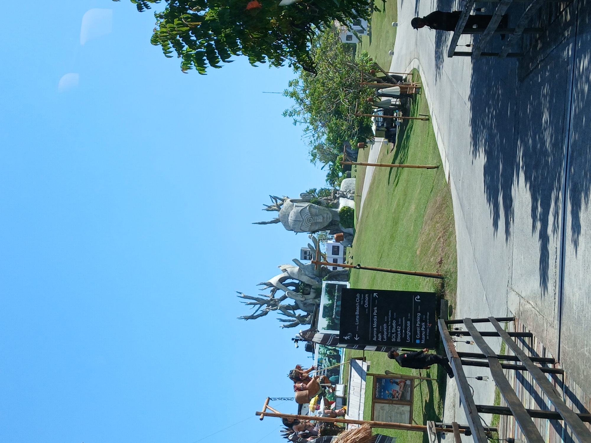 In Mengwi, Indonesia, on a sunny day, a directional signpost stands on a paved pathway. The signpost lists various locations and their distances, including "Luna Beach Club," "Aurora Media Park," "SOL Studio," "Block42," "Longhouse," and "Nuonu Park." A man in a dark uniform walks away from the signpost, while another man in a similar uniform stands near a wooden railing. In the background, a large-scale artistic sculpture depicting a human face with intricate carvings is visible, along with other abstract sculptures and lush green grass. A road curves through the landscape, leading towards buildings and trees.