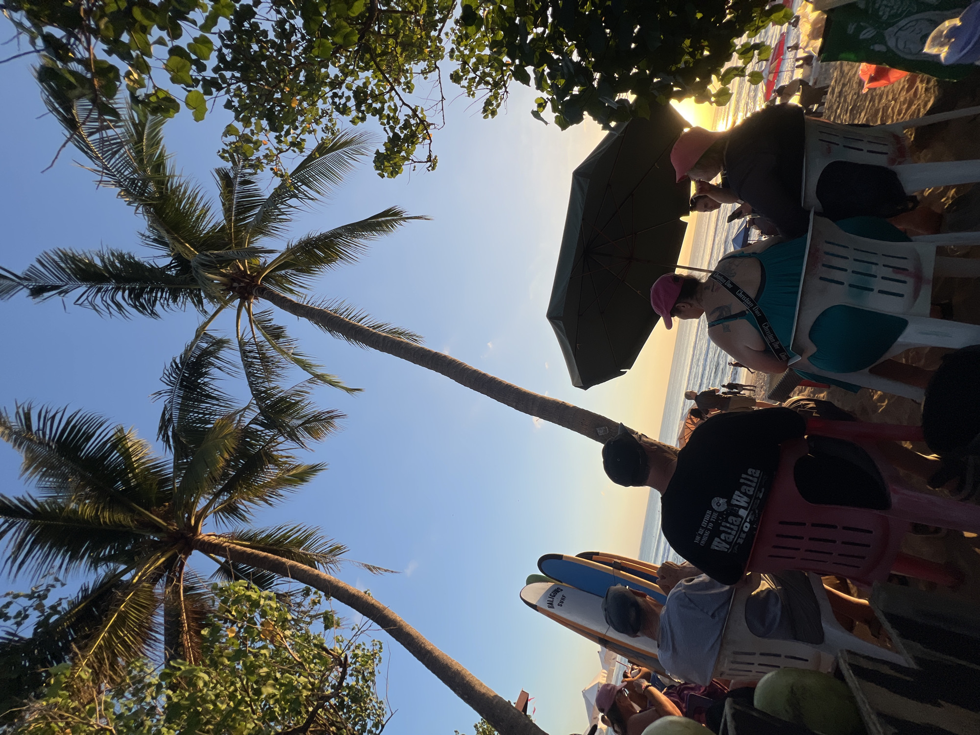 In Kuta, Indonesia, at sunset, several people are seen sitting on chairs on a beach, facing the ocean. Two prominent palm trees frame the scene, their fronds reaching towards the clear blue sky. In the foreground, a group of people are seated, some looking out at the water. One person in the center is wearing a black t-shirt with white lettering that says "YOU'RE EITHER COMING TO THE / WALLA WALLA". To their left, two surfboards stand upright, one with the word "BALIGUNS" visible. In the background, the sun is setting over the ocean, casting a warm glow on the scene. Other beachgoers, umbrellas, and some tents are visible further down the beach, suggesting a lively atmosphere. The sand is a light brown, and the water appears calm.