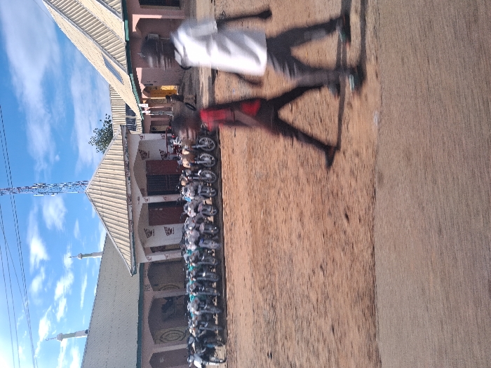 The image displays an urban street scene in Beli, Nigeria, under a blue sky with scattered white clouds. The foreground consists of a concrete strip transitioning into a wide expanse of reddish-brown dirt. Two individuals are captured in motion blur, walking from right to left across the dirt; the person on the left wears a dark top and dark pants, while the person on the right wears a white top and dark pants.
In the mid-ground, a line of approximately 20-30 motorcycles, mostly silver and white, are parked directly in front of a single-story building. This building features a light-colored pitched roof, arched entrances, and "Honda" logos affixed to its facade. To the left and slightly behind this structure is a larger building with a similar light-colored pitched roof and two minarets, suggestive of a mosque, displaying a circular design on its visible wall. To the right of the central building, another single-story building with a light-colored pitched roof and reddish-brown walls is visible, with a faint human figure in one of its doorways. A tall, lattice-style communication tower with multiple antennas stands prominently in the background, positioned to the right of a visible green tree. Overhead wires are visible across the top of the frame.