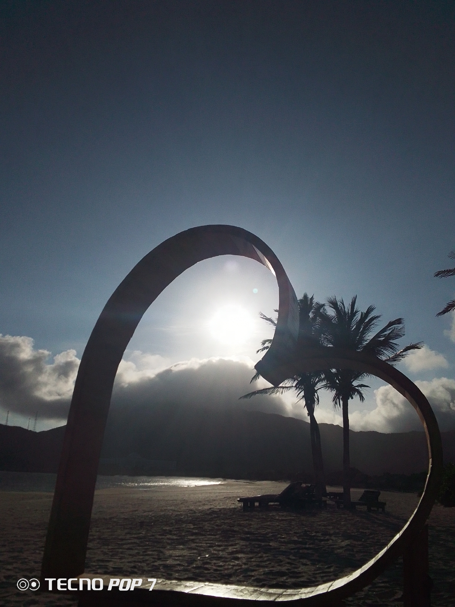 A low-angle shot depicts a beach scene at Pedro González, Venezuela. A heart-shaped metal structure dominates the foreground. The bright sun is partially obscured by clouds and mountains in the background. Two palm trees stand behind the heart. Three lounge chairs are on the sand between the structure and trees. "TECNO POP 7" is in the lower left corner.