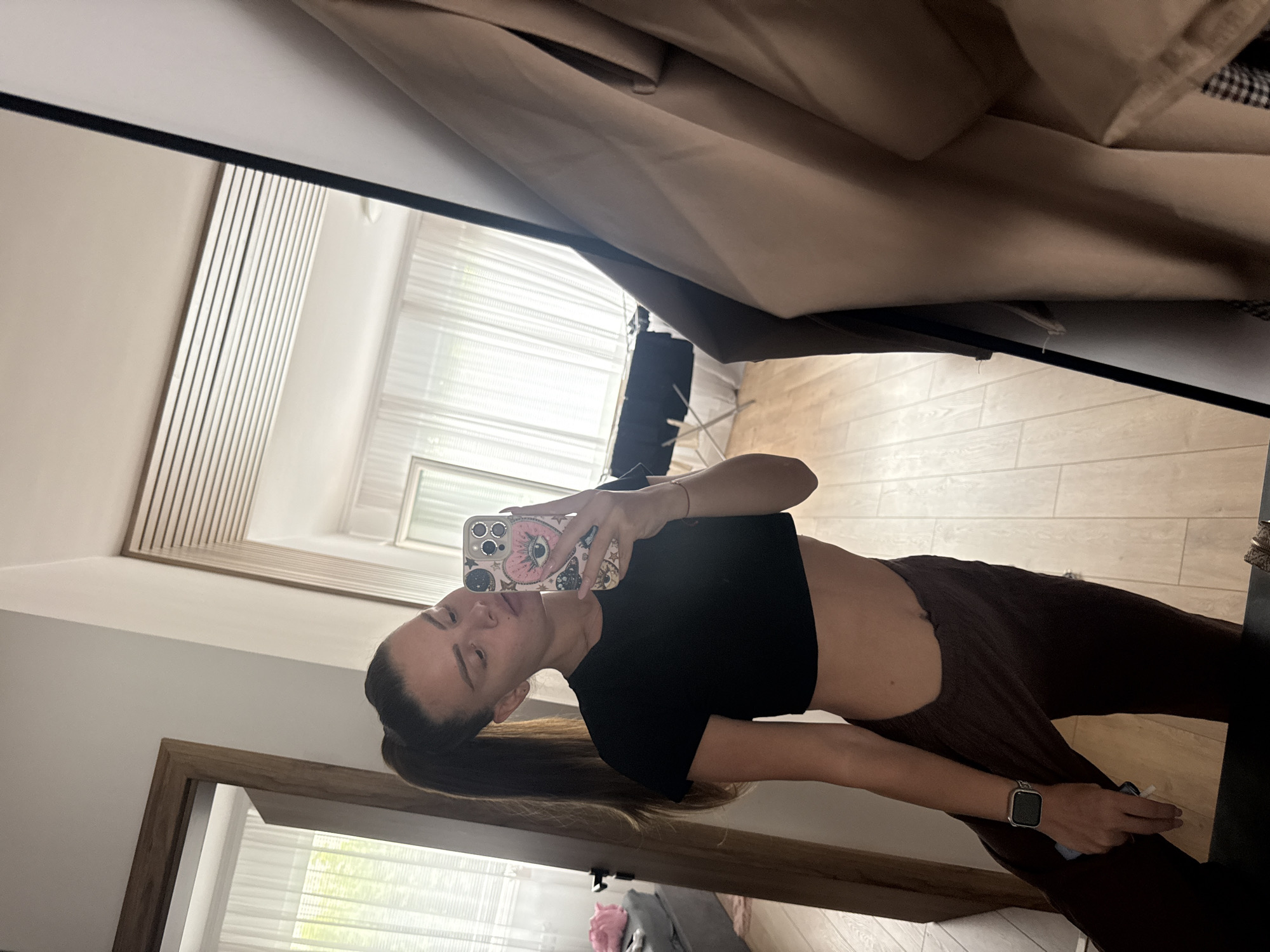 In Plovdiv, Bulgaria, a young woman with her hair in a high ponytail is captured in a mirror selfie. She is wearing a black crop top and dark brown pants that sit low on her hips, revealing her midriff. On her left wrist, she wears a smartwatch. Her right hand holds a smartphone with a decorative case featuring a pink eye design. The background, visible in the reflection, suggests an indoor setting with wooden flooring, white sheer curtains, and modern ceiling paneling. To the right of the woman, a tan coat is draped over a closet door. The overall impression is of a casual, personal moment captured in a contemporary living space.