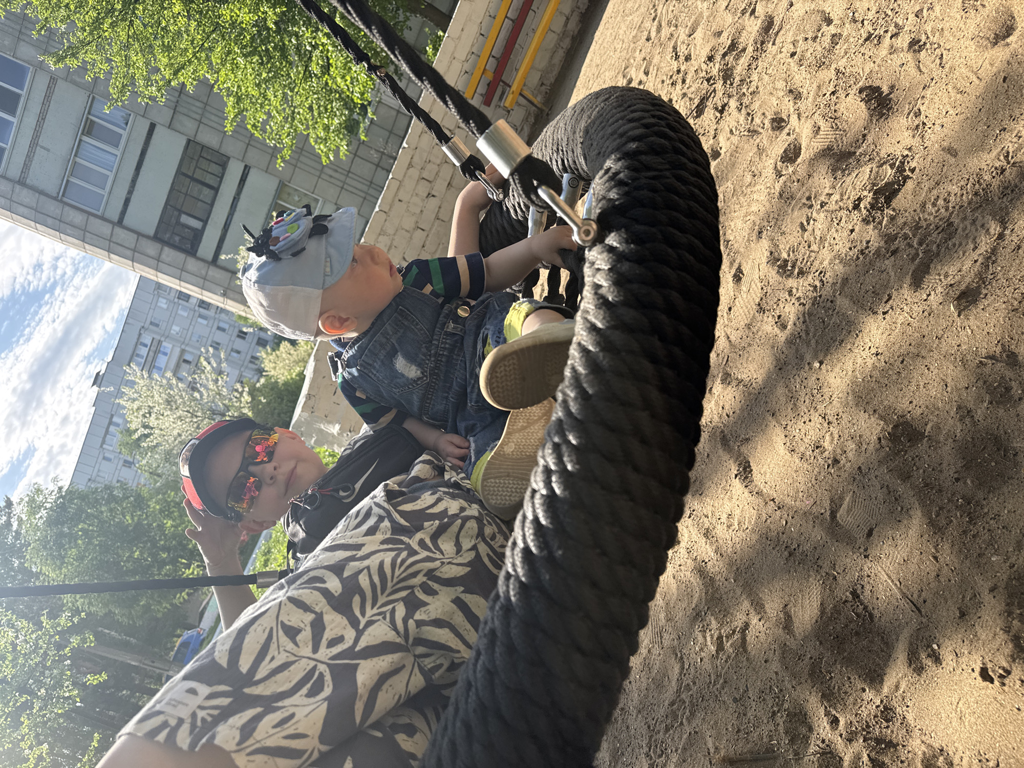 A photograph depicts two individuals in a rope swing. The younger child, wearing a denim overall and a light-colored cap with a decorative emblem, sits in the swing, holding onto the rope. An older child, sporting sunglasses and a patterned shirt, is positioned behind the younger one. The setting appears to be a playground, with a sand-covered ground visible beneath the swing. A residential building is visible in the background, surrounded by trees. The location is the city of Chaikovsky, Russia.