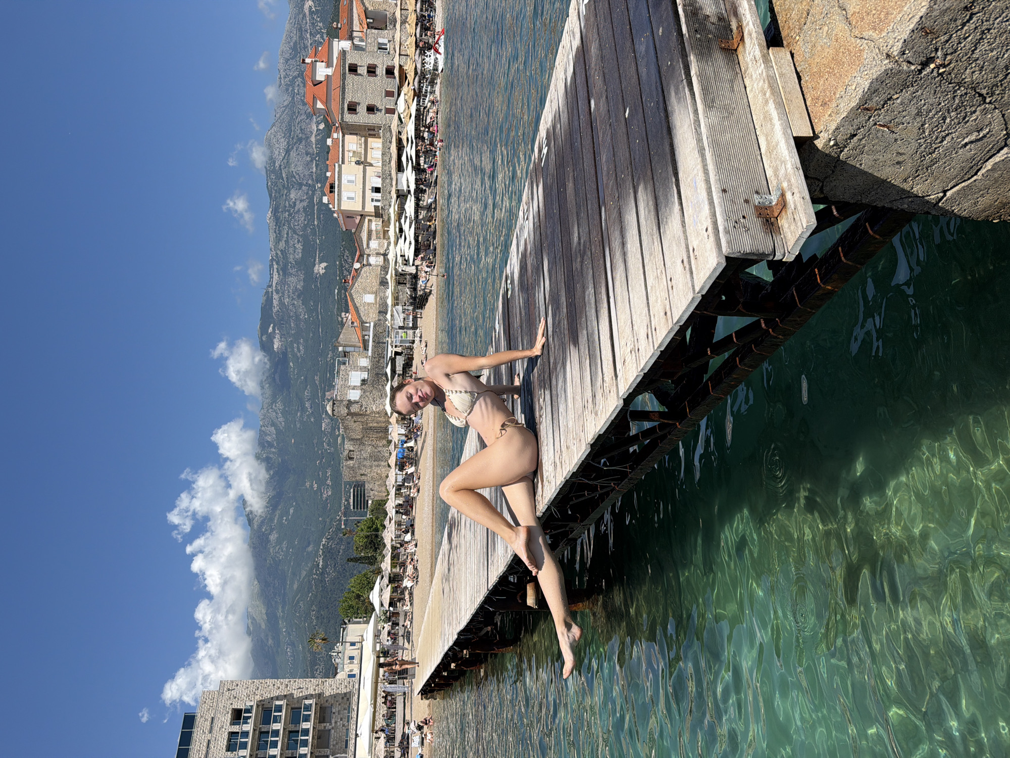 A young woman in a beige bikini is sitting on a wooden pier with her legs outstretched. She is looking at the camera with a slight smile. The pier extends into clear, turquoise water. In the background, there is a beach with umbrellas and people, and beyond that, buildings and mountains. The sky is blue with some white clouds. The scene appears to be the coast of Budva, Montenegro, known for its beaches and old town.