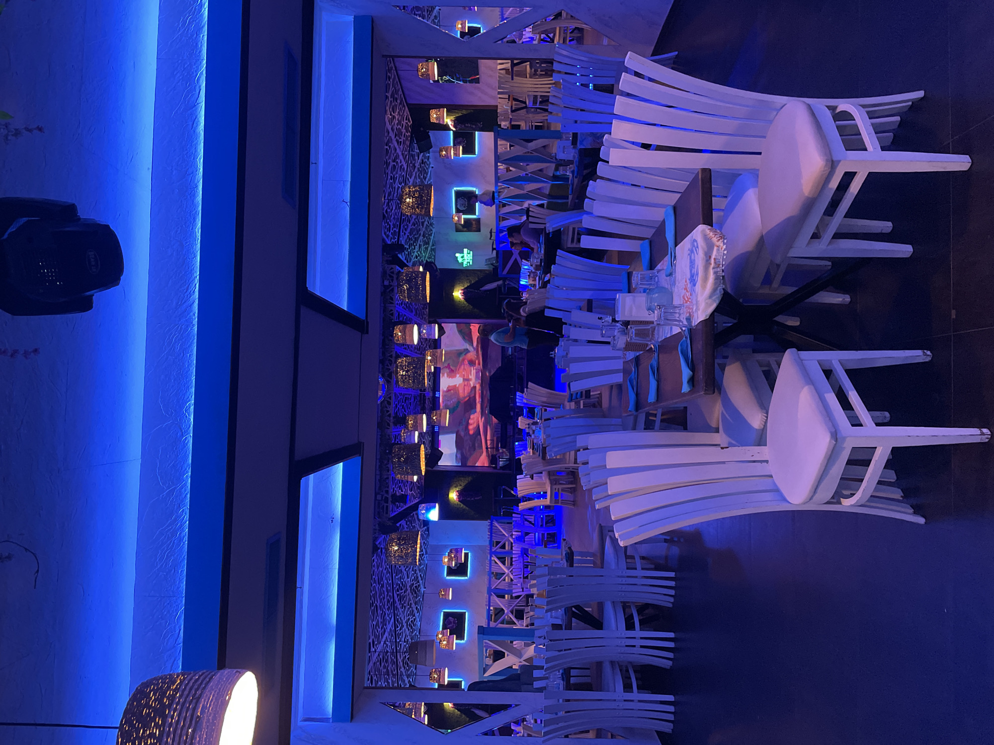 The image depicts the interior of a modern restaurant, bar, or lounge in Plovdiv, Bulgaria, characterized by a predominant cool blue ambient lighting scheme. The setting appears to be during the evening or night, relying entirely on artificial light.
The space is illuminated by numerous blue LED strip lights integrated into the textured walls and ceiling panels, creating a vibrant and somewhat futuristic atmosphere. This strong blue light is softened by the warm, orange glow emanating from several decorative pendant lamps with woven shades, which are suspended throughout the room.
The venue is furnished with numerous white, contemporary-style chairs, some with light-colored cushions, paired with dark tables. Many of these chairs are stacked neatly on tables, suggesting that the establishment might be preparing for opening or closing, or perhaps has sections that are currently not in active use. Tables are set with clear glassware and blue napkins.
Decorative elements include both textured wall surfaces highlighted by the blue LEDs and intricate geometric latticework on other wall sections. In the background, multiple large screens display colorful imagery, possibly scenic landscapes or abstract art, adding to the visual ambiance. A black stage lighting fixture, with the word "BEAM" partially visible, is mounted on the far left, hinting at potential entertainment or events.
A few patrons are visible seated at tables in the mid-ground, suggesting that the establishment is open and serving guests, who appear to be dining or socializing in this chic and relaxed environment.