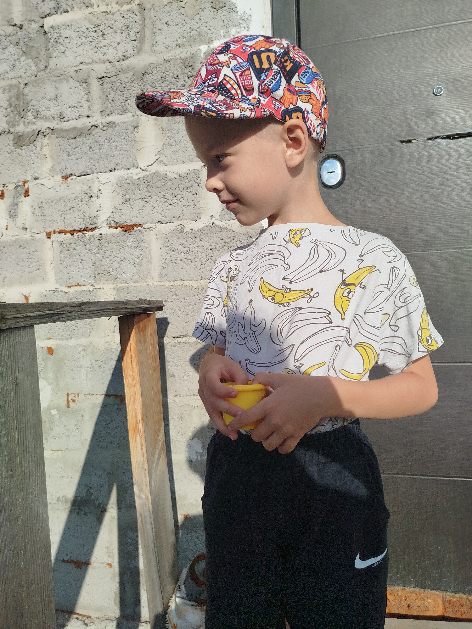 A young boy wearing a colorful, patterned baseball cap and a white t-shirt with a banana print is standing outdoors. He is holding a small yellow cup in his hands. The background consists of a rough, light-colored brick wall on the left and a dark metal door on the right. Wooden planks are visible against the brick wall, casting shadows. The scene appears to be in a residential or outdoor area.