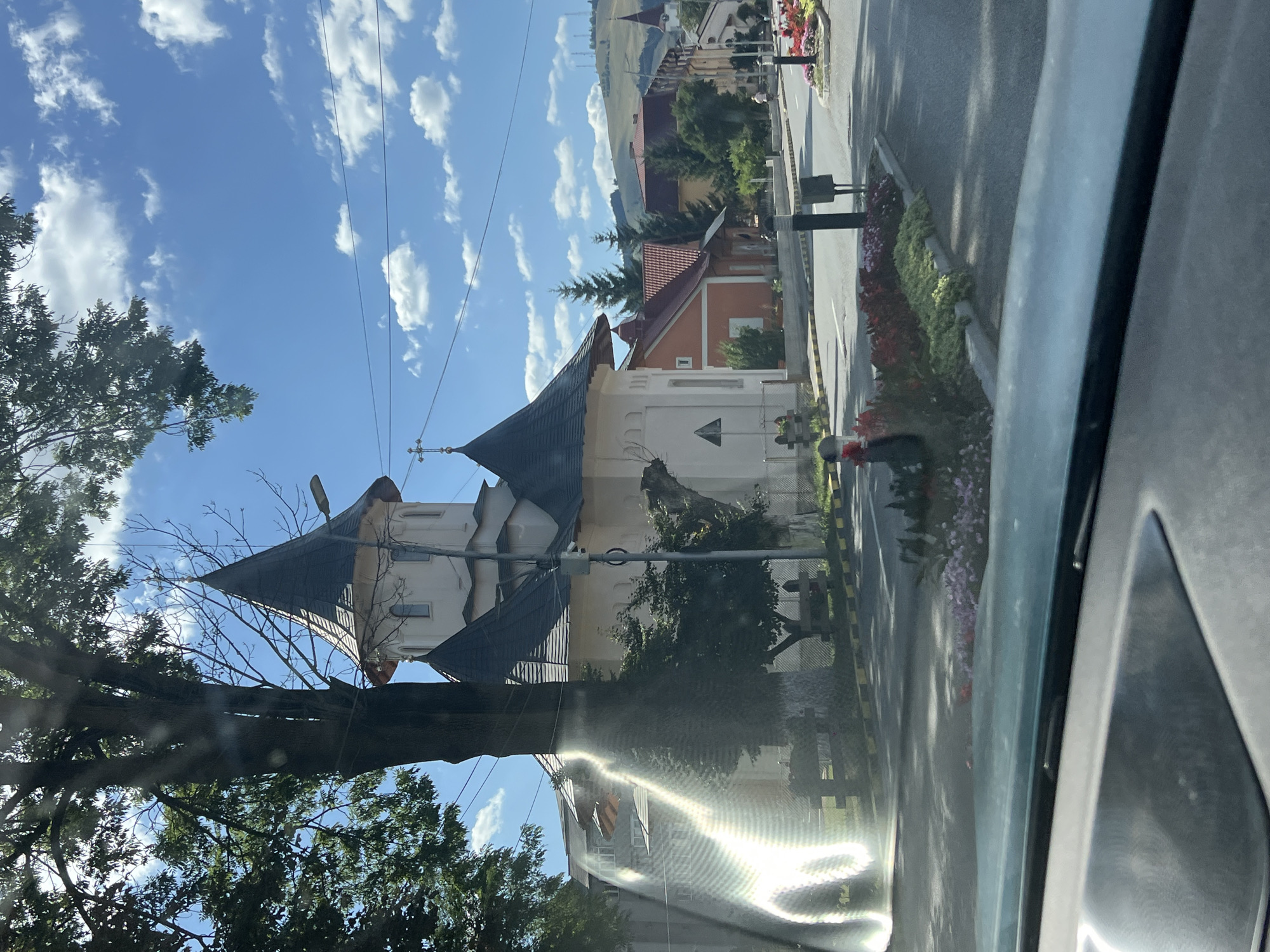 The image, captured from inside a vehicle, shows a street scene in Gura Humorului, Romania, on a sunny day with scattered clouds. In the foreground, the car's dashboard is visible. The middle ground features a road with a tree on the left and a building with a prominent, conical-shaped roof, likely a church, on the right. The building is light-colored with dark roofing and a cross atop the spire. Adjacent to the road, there are flower beds with red and pink flowers. Further down the street, houses with red roofs and other buildings can be seen, leading towards a hilly landscape in the background. The overall atmosphere is peaceful and urban, with elements of nature and architecture harmoniously present.