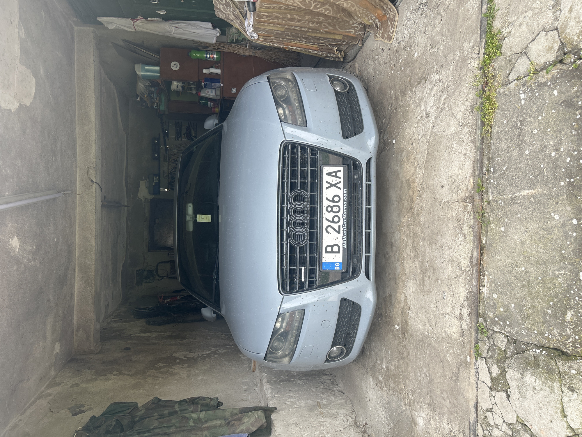 The image shows a gray Audi car parked inside what appears to be a garage or storage space. The garage has concrete walls and floor. The car's license plate, reading "B 2686 XA," is visible. The area outside the garage shows cracked concrete and some vegetation growing along the edges. The environment is cluttered, with various items stored on shelves inside the garage. The location is Unknown, Unknown. There is no indication of any people in the image.