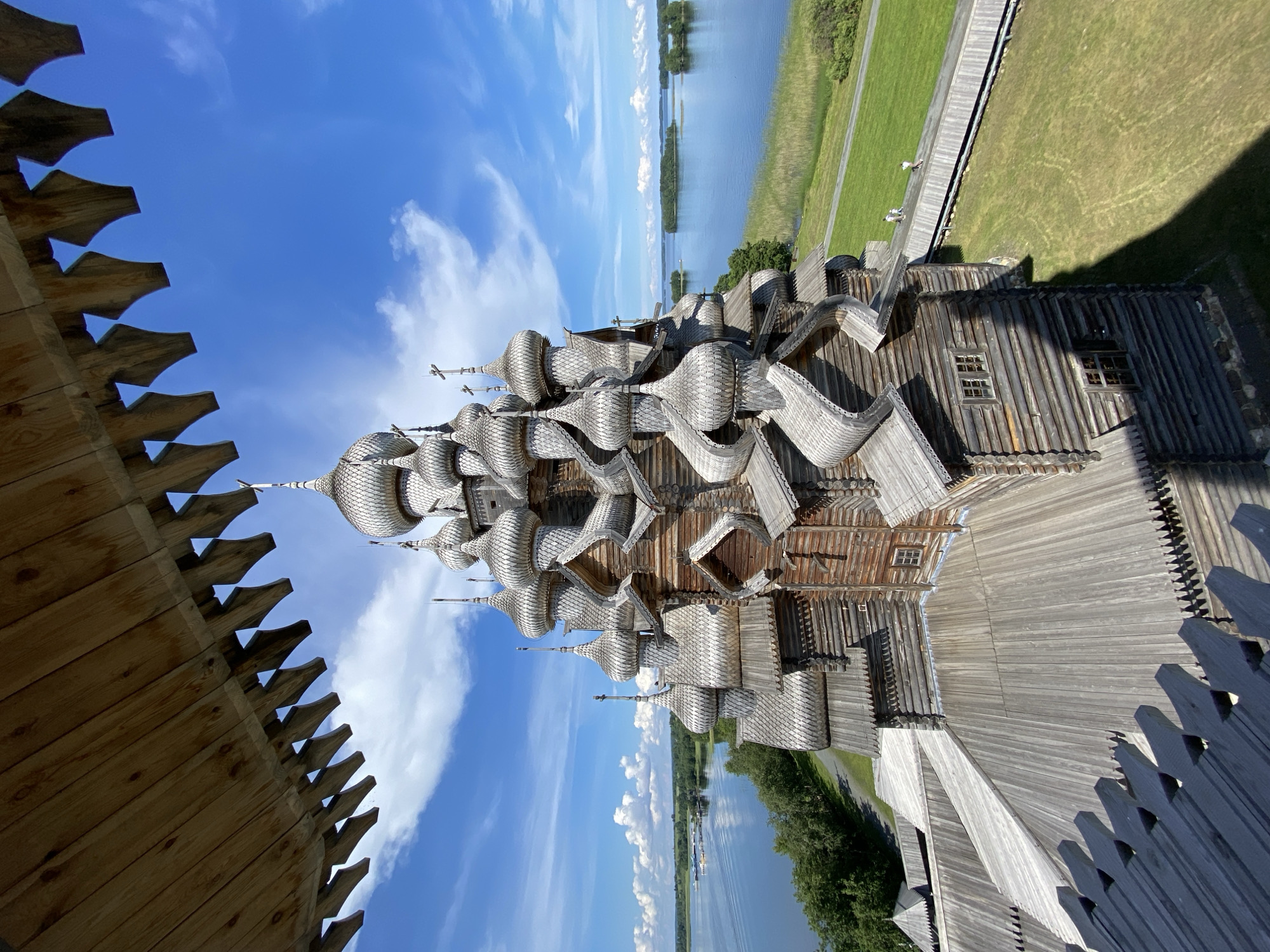 The image displays a detailed view of the Kizhi Pogost, a historical architectural ensemble located on Kizhi Island in Karelia, Russia, near the city of Boyarshchina. The primary subject is the Church of the Transfiguration, a magnificent wooden church characterized by its numerous domes and intricate wooden construction. The church is situated within a grassy, green landscape adjacent to a body of water, likely Lake Onega, with islands visible in the distance. The sky is a bright blue with scattered white clouds. In the foreground, there is a wooden structure, possibly a viewing platform or part of another building, with distinctive carved wooden elements. Two individuals are visible walking on a wooden pathway to the right of the church, suggesting the area is a tourist or historical site. The image is taken from an elevated perspective, looking down and across the church.