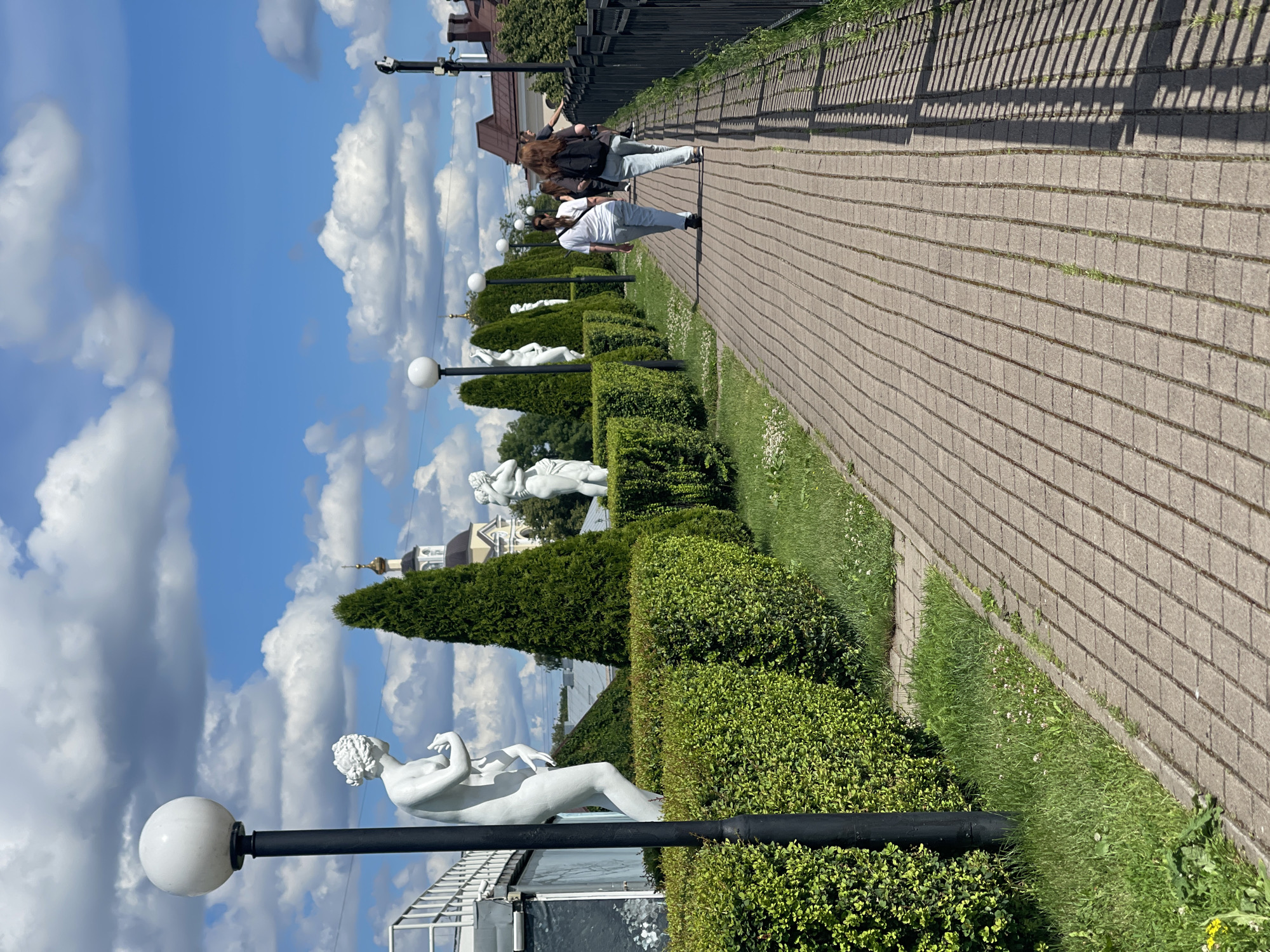 The image depicts a sunlit pathway in Vyborg, Russia, lined with manicured hedges and classical white sculptures. The pathway, paved with rectangular bricks, stretches into the distance, bordered by lush green grass and trees on one side and a dark metal fence on the other. Several individuals, dressed casually in t-shirts and jeans, walk along the path, facing away from the camera. In the background, a church with a golden dome is visible, along with residential buildings and a bright blue sky dotted with white, fluffy clouds.
