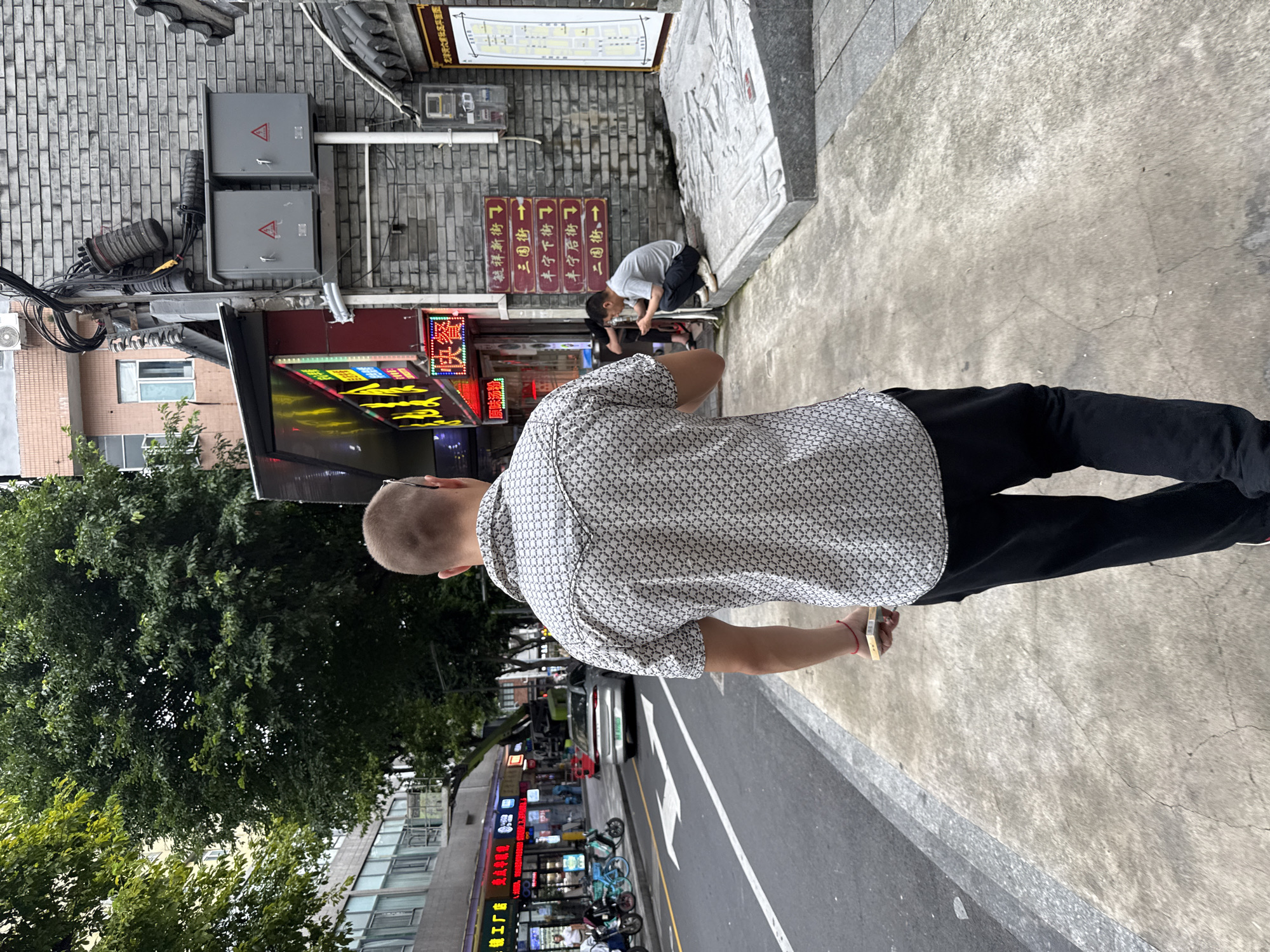 A man walks away from the camera down a sidewalk in Liwan District, China. He is wearing a short-sleeved patterned shirt and black pants. In his right hand, he holds a small stack of bills with a red string tied around it. To his left, a street runs alongside the sidewalk with parked motorcycles, a silver car, and storefronts. To his right, a brick building with electrical boxes and signage stands adjacent to the sidewalk. A second man is seated on a raised platform in front of the brick building, looking at his phone. Overhead, trees and other buildings are visible. The lighting suggests it is daytime.