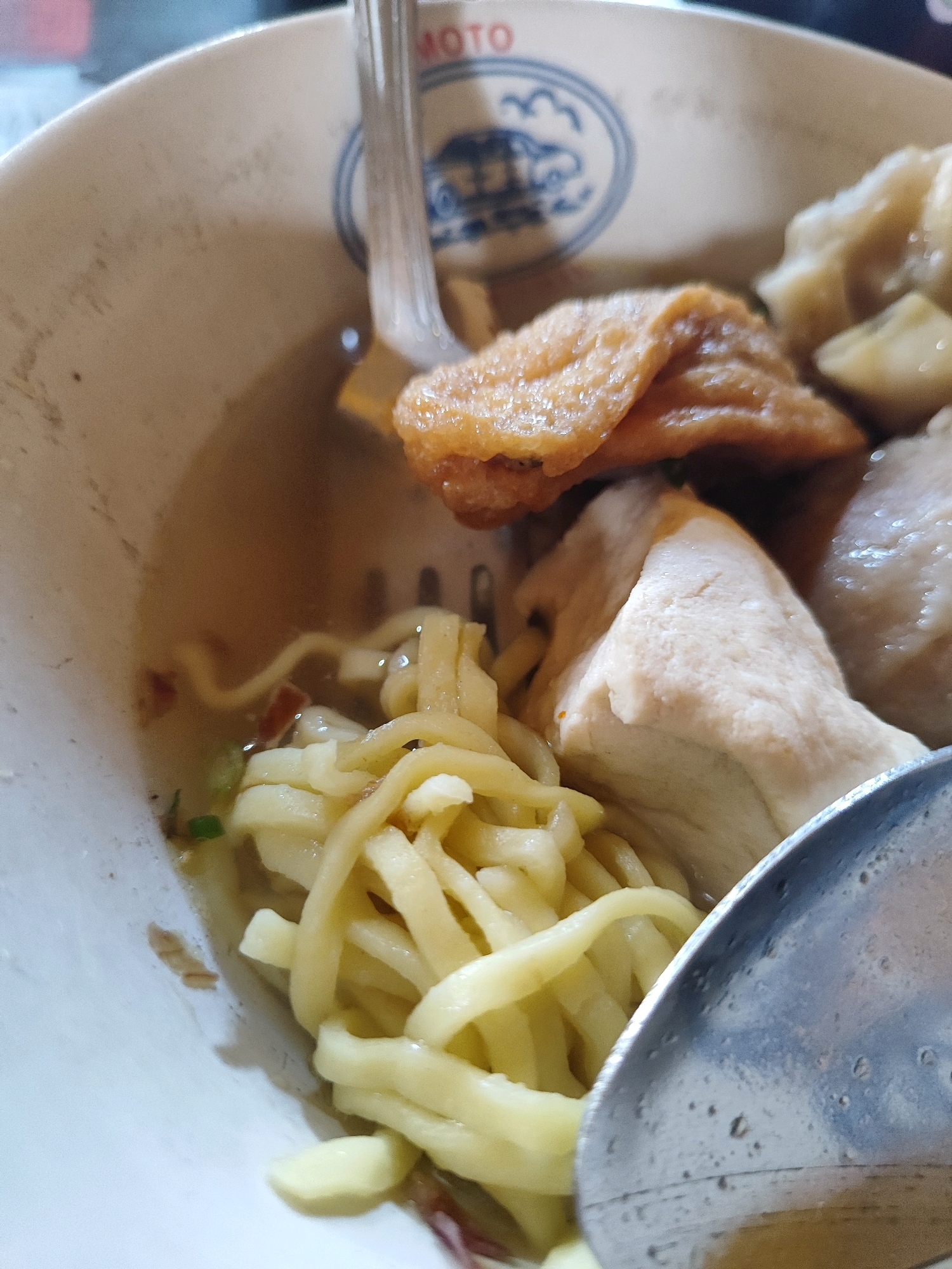 A close-up, eye-level shot captures a bowl of Indonesian cuisine, likely bakso or a similar noodle soup, presented in a white ceramic bowl with blue trim that includes the word "MOTO". The bowl contains a light broth, thick yellow noodles, a triangular piece of pale tofu, and what appear to be fried wonton or dumpling wrappers. A silver spoon, partially submerged, is visible in the lower right quadrant. The background is blurred, suggesting an indoor setting. The image is cropped to focus on the contents of the bowl, with no people or external environment clearly visible. This scene is characteristic of a local eatery in Bondowoso, Indonesia, where such dishes are commonly served.