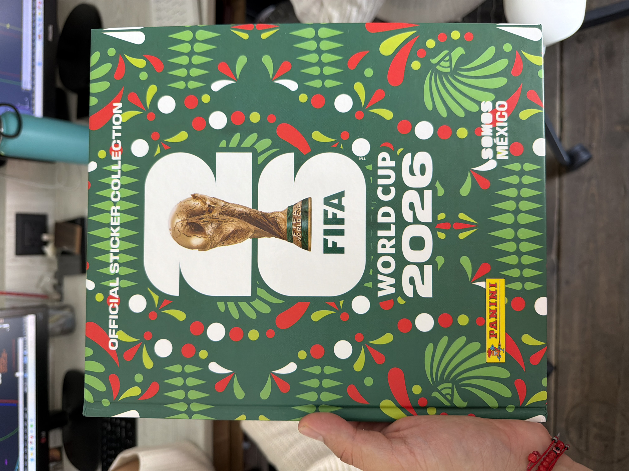 The image shows a FIFA World Cup 2026 official sticker collection album. The album has a green background with red, white, and green decorative patterns. The text "OFFICIAL STICKER COLLECTION", "FIFA WORLD CUP 2026", and "Somos Mexico" are visible. The album also features a stylized image of the FIFA World Cup trophy. The location of the image cannot be determined to be in Puebla, Mexico, based on the visual information provided. The album is being held by a person. The background shows a desk with monitors and other items.