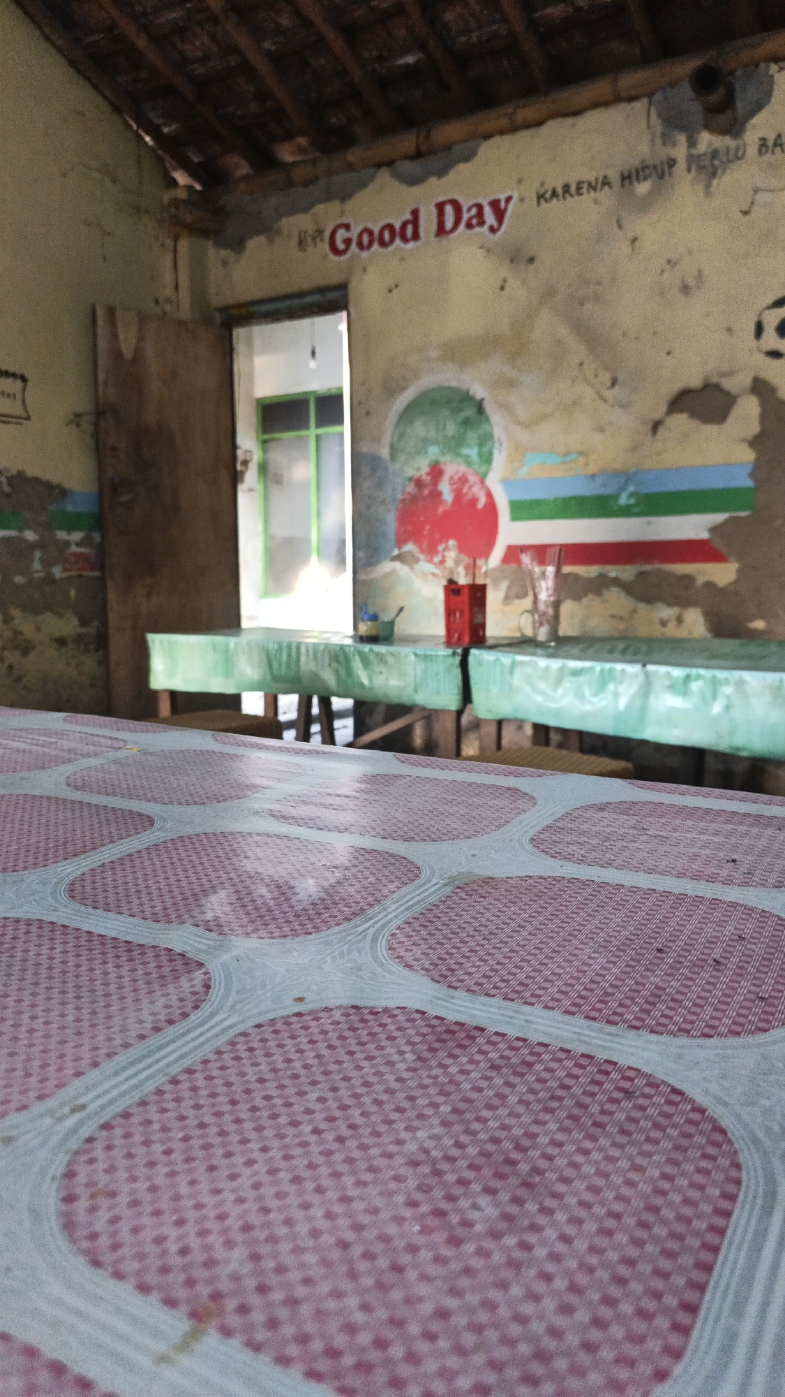 The image depicts a rustic, open-air eatery in Dawe, Indonesia, characterized by its weathered walls and simple furnishings. A wooden door stands ajar, revealing a glimpse of an interior with green-framed windows. The walls are adorned with faded paint, featuring a prominent "Good Day" slogan in red lettering and a series of colorful stripes. Two long tables, covered with green plastic sheeting, are set up, suggesting a communal dining area. On one table, a red cigarette box and a glass are visible. The foreground is dominated by a table covered in a pink and white patterned plastic cloth, its texture creating a visually engaging foreground element. The overall impression is one of a humble, local establishment, likely a warung, a common type of small eatery in Indonesia, in the rural district of Dawe, situated in the Kudus Regency of Central Java.