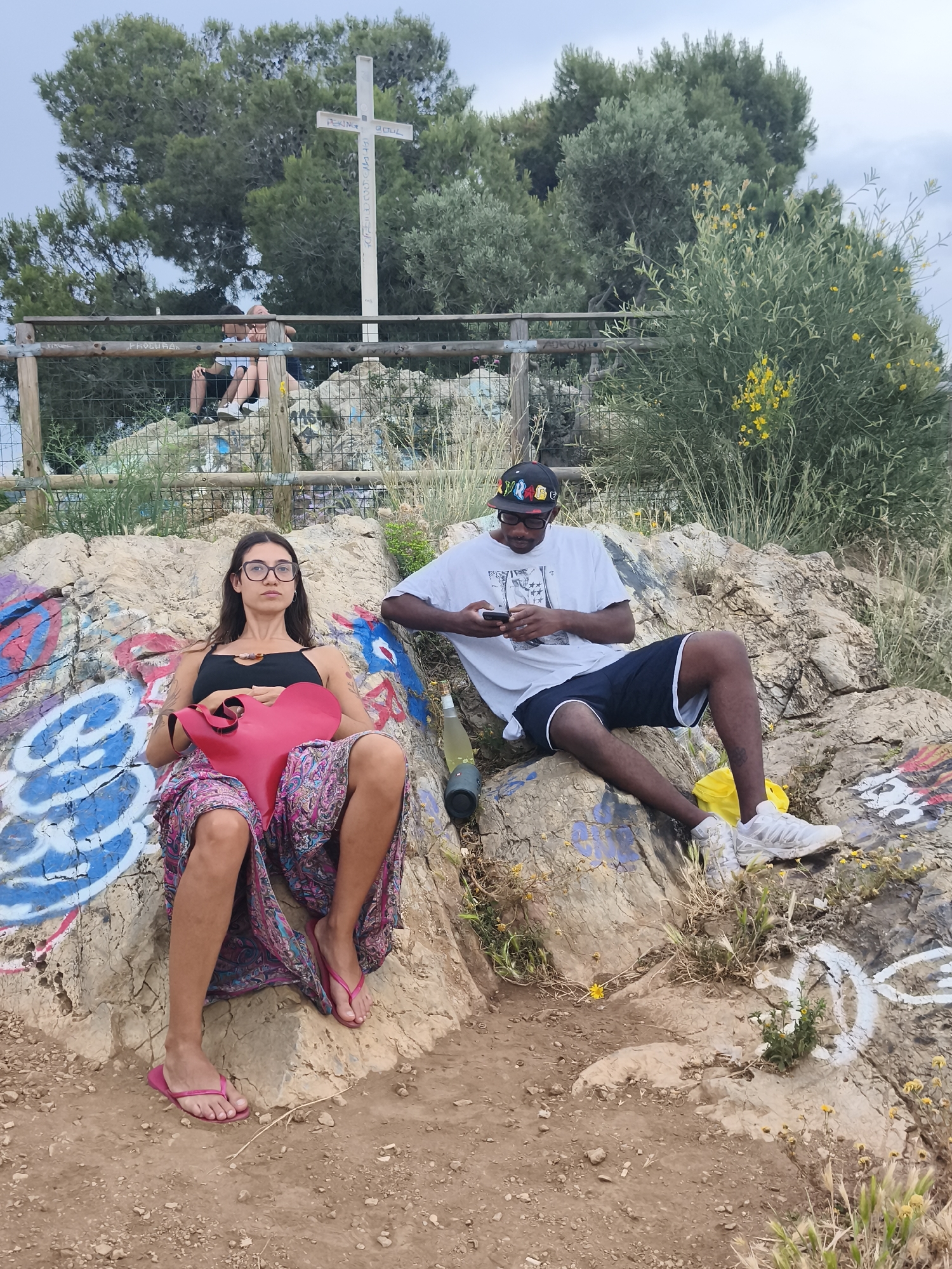 The image shows two individuals seated on a rock formation that has been decorated with graffiti. The woman on the left is wearing a black top, a long, patterned skirt, and pink flip-flops. She is holding a red, heart-shaped object. The man on the right is wearing a white t-shirt, black shorts, white sneakers, and a cap with colorful writing. He is looking at his phone. There is a bottle lying near him.
In the background, a wooden fence and green vegetation are visible, as well as a white cross on top of a hill. The cross has graffiti written on it. Two people are sitting on the other side of the fence, near the cross.
The image was taken at Bunkers del Carmel, a former anti-aircraft battery site in Barcelona, Spain.