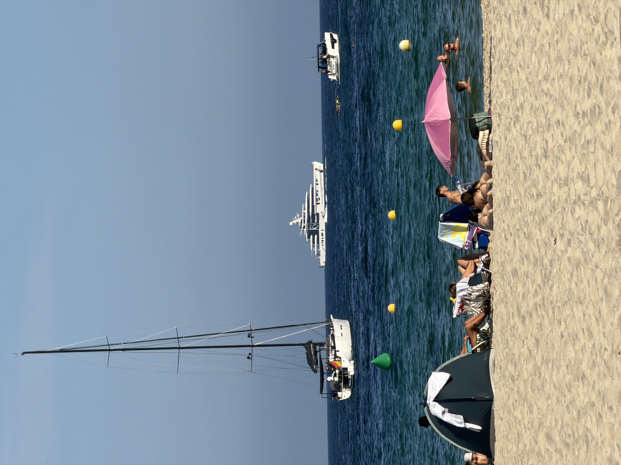 The image depicts a sunny day at the beach in Calvià, Spain, with several sailboats and yachts in the background. In the foreground, people are relaxing on the sandy beach. Some are seated in chairs, while others are sunbathing. A blue tent is set up, and a pink umbrella provides shade. The sea is a vibrant turquoise color, with a few buoys floating on the surface. A large white yacht is visible in the distance, along with a smaller white boat. The sky is clear and blue.
