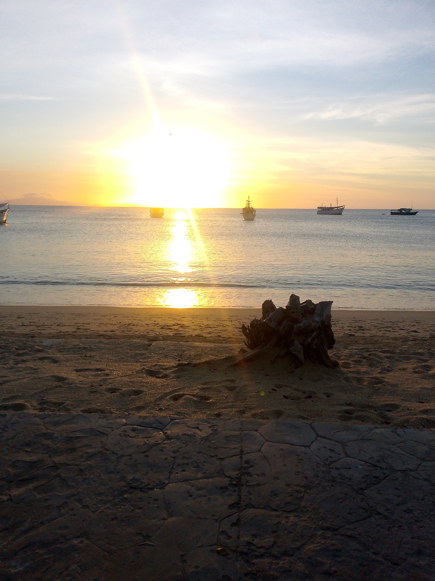 The image displays a coastal scene at either sunrise or sunset in Juan Griego, Venezuela. A bright sun is positioned low on the horizon, slightly left of the frame's center, emitting a strong light that reflects across the calm water in a shimmering, golden path. The sky above transitions from light blue with faint clouds at the top to a vibrant yellow and orange gradient near the horizon. A small dark bird is faintly visible in the upper central sky.
On the water, several boats are present: a portion of a vessel on the far left, a light-colored boat with a mast structure on the left, a darker boat with a mast further back towards the center, and a smaller, dark boat on the far right.
The immediate foreground consists of a sandy beach exhibiting footprints and an uneven texture. A large, dark, gnarled object, resembling a tree stump or significant driftwood, is centrally located on the sand. In the very bottom foreground, a dark, heavily shadowed paved surface with an irregular, possibly hexagonal, pattern is visible directly in front of the sand. No people are present, and no specific actions or interactions are depicted beyond the environmental elements.