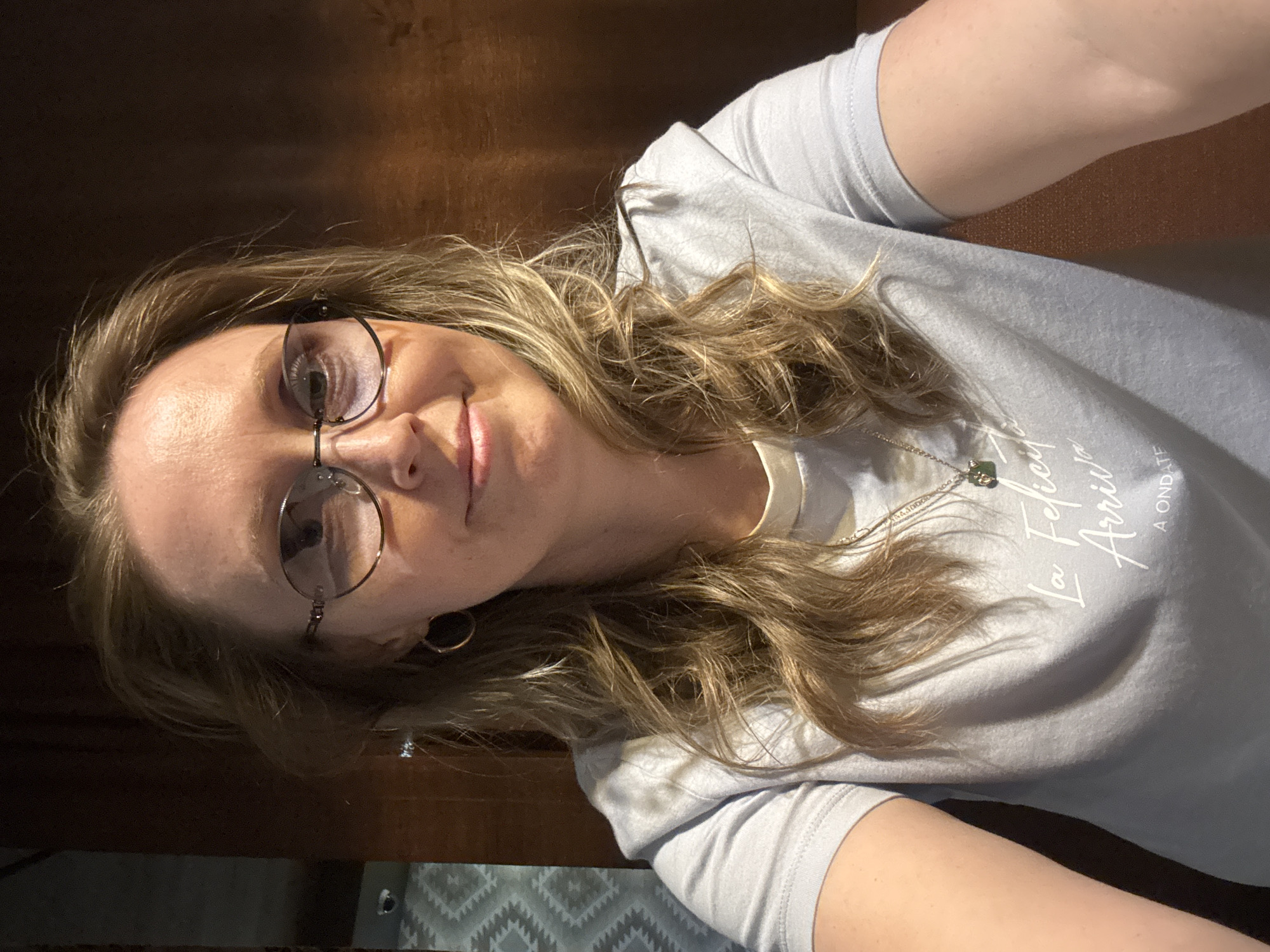 A woman with blonde, wavy hair and glasses is looking at the camera. She is wearing a light-colored t-shirt and a necklace with a small pendant. The background appears to be a wooden surface and part of a patterned blanket or throw. It is not possible to determine the location within Balezino, Russia, from the image.