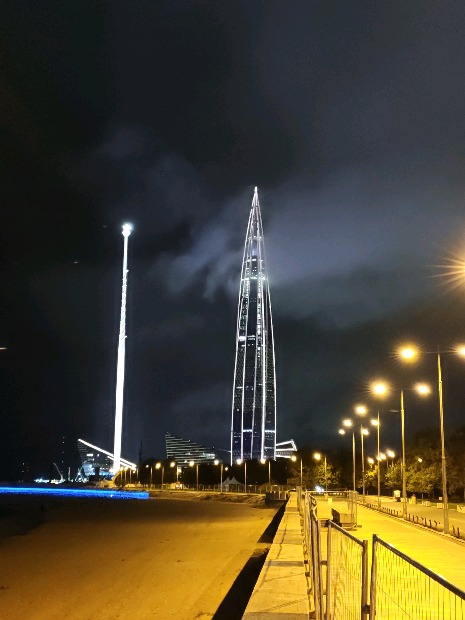The image depicts a nighttime scene in Saint Petersburg, Russia, focusing on the Lakhta Center skyscraper and a tall, illuminated tower. The Lakhta Center, a striking modern skyscraper with a twisted glass facade, dominates the right side of the frame, its lights outlining its distinctive shape against the dark sky. To the left, a tall, slender tower, possibly a construction crane or a decorative illuminated structure, glows brightly with white light.
In the foreground, a paved area, possibly a promenade or walkway, is illuminated by warm yellow streetlights. A yellow metal safety fence runs along the edge of the walkway, separating it from what appears to be a lower, sandy or unpaved area. Trees line the street in the background, their silhouettes visible against the city lights. A body of water or a brightly lit area with blue lights is visible in the distance on the left, hinting at a waterfront location. The overall atmosphere is urban and modern, with a focus on impressive architecture and artificial illumination. No people are visible in the scene.