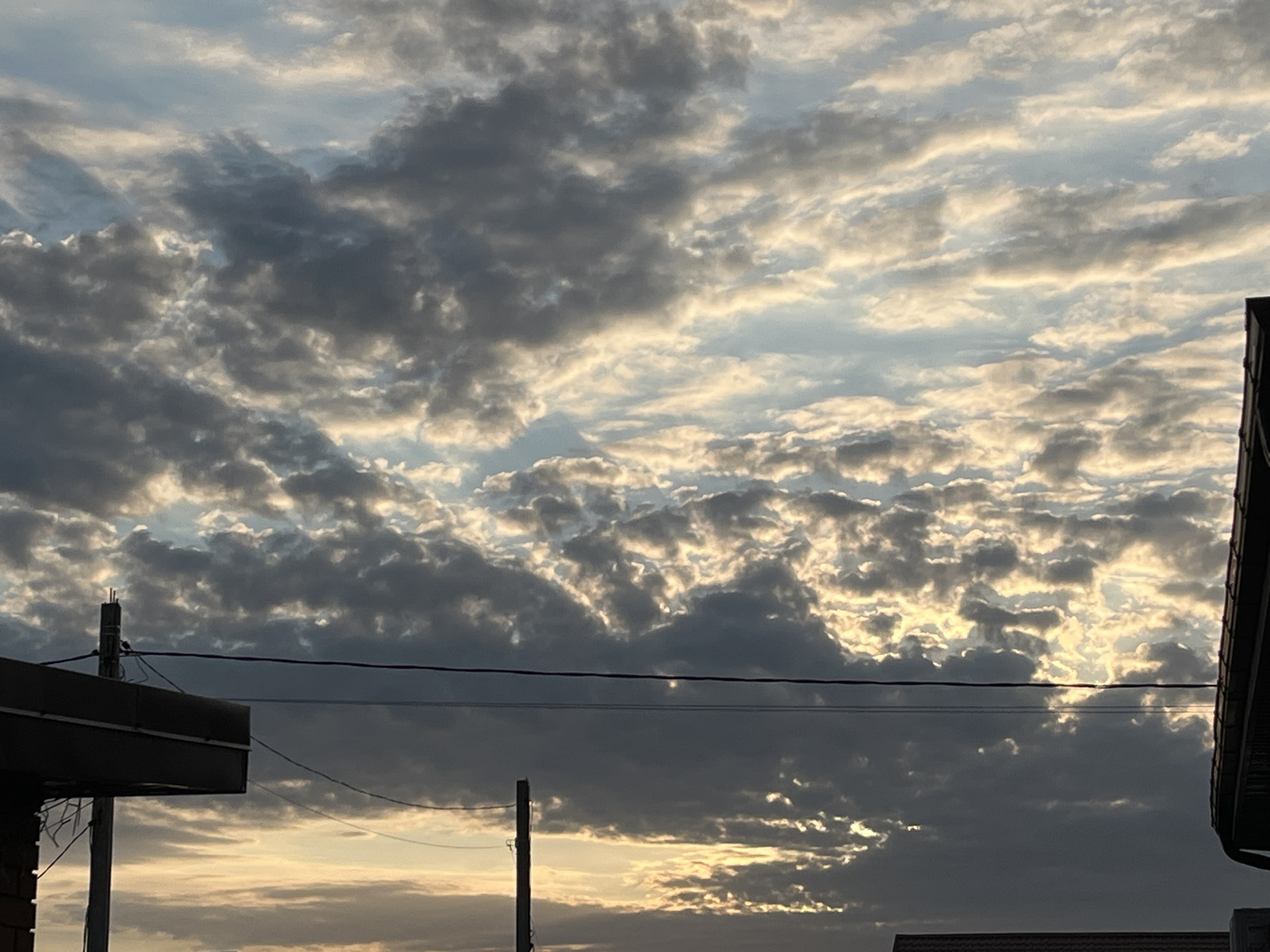 The image shows a cloudy sky at what appears to be sunset or sunrise. The clouds are a mix of dark grey and lighter, illuminated areas, suggesting the sun is behind them or breaking through. In the foreground, silhouetted against the sky, are parts of buildings and utility poles with wires. On the left, a structure with a dark overhang and a brick base is visible, with a utility pole in front of it. Another utility pole is positioned further to the right. The environment appears to be urban or suburban, based on the man-made structures. The time of day is indicated by the low angle of the light and the warm tones in the sky.