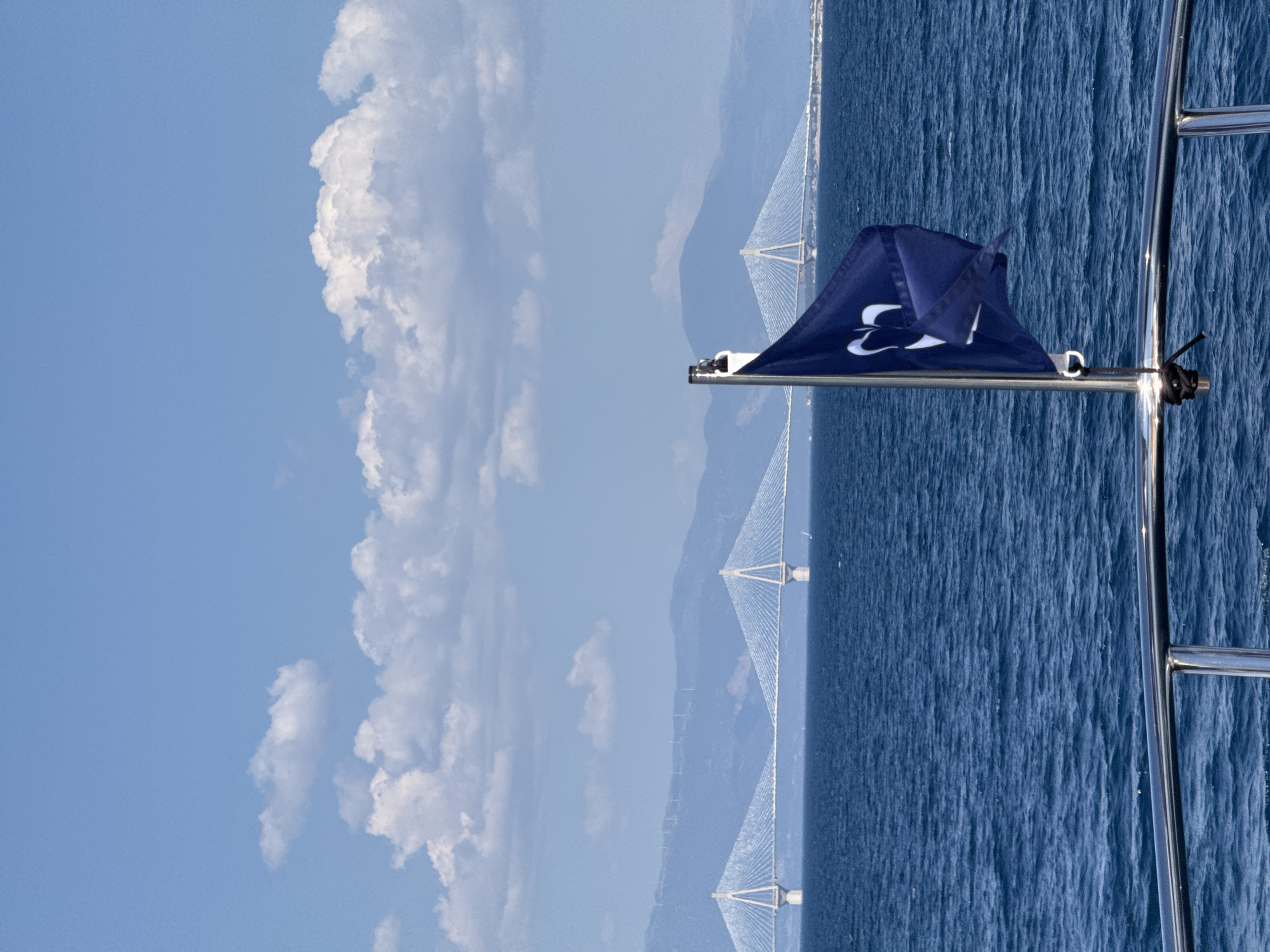 The image captures a scenic view of the Rio-Antirrio Bridge in Patras, Greece, from the perspective of a boat. The vast expanse of deep blue ocean dominates the right side of the frame, with gentle ripples suggesting a calm day. In the distance, the impressive cable-stayed bridge spans across the water, its white towers and cables stark against the hazy blue sky and distant mountains. The sky on the left is a bright, clear blue, punctuated by large, fluffy white clouds that cast soft shadows.
In the foreground, a dark blue flag with a white logo is affixed to a metal pole. The logo appears to be a stylized "3" or a similar abstract design. A metal railing runs along the right edge of the image, hinting at the boat's deck. A black rope is tied to the railing. The overall impression is one of a beautiful, sunny day, likely late morning or early afternoon, given the brightness of the sky. The scene evokes a sense of peace and the enjoyment of a maritime journey with a prominent landmark in the background. There is no visible text other than the logo on the flag.