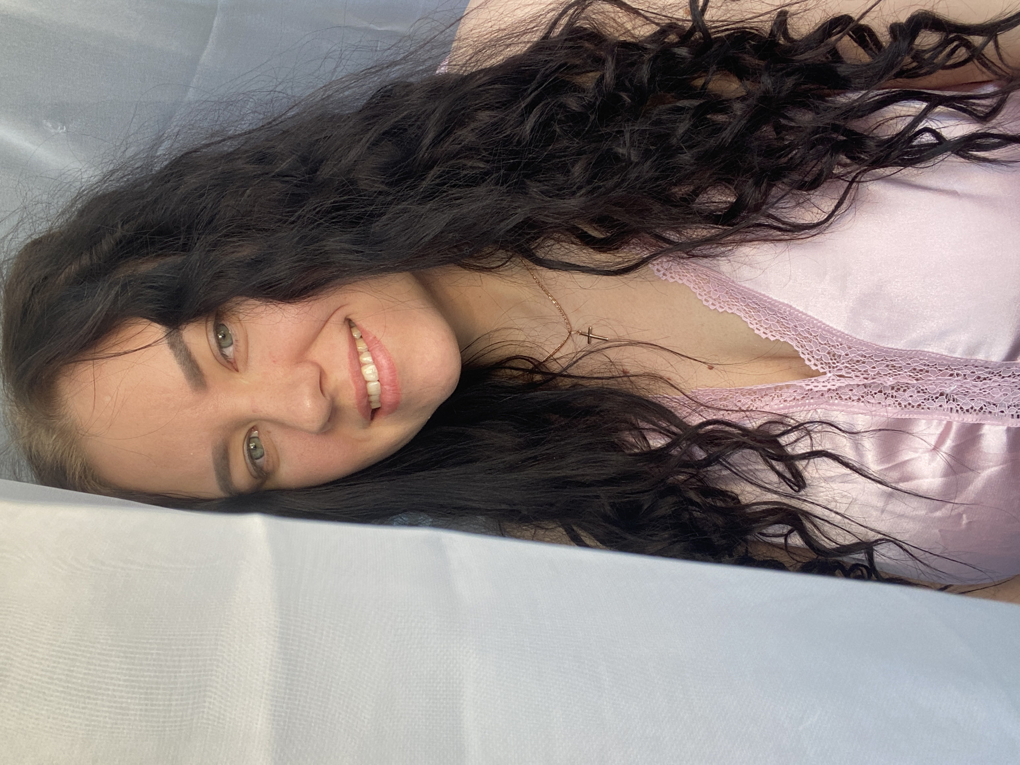 A young woman with long, dark, curly hair is lying on a white surface, possibly a bed or examination table. She is wearing a light pink top with lace trim around the neckline. She is smiling, and her head is turned slightly to the side. She has fair skin and visible eyebrows. A thin gold necklace with a cross pendant is around her neck. The background appears to be a plain, light-colored fabric. There's no explicit indication of Chaikovsky, Russia in the image.