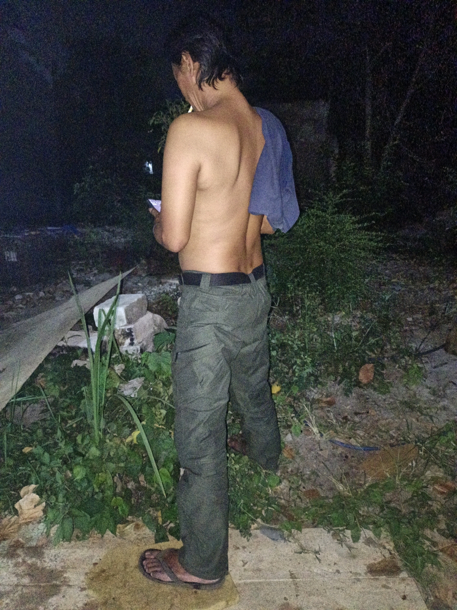 A shirtless person, with dark hair and fair skin, stands facing away from the camera in a dimly lit, outdoor environment. They are wearing dark, tactical-style pants with a black belt and a blue cloth draped over their left shoulder. Their right hand holds a mobile device, and they appear to be looking at it. The person is standing on a light-colored, wet surface that transitions to dirt and vegetation. In the background, there are dense green bushes and trees, suggesting a natural or semi-rural setting. A large, light-colored, angular object, possibly construction material, lies on the ground to the left of the person, along with some bricks. The lighting suggests it is nighttime or very early morning. The location is Cikal, Indonesia, a rural area known for its natural landscapes.
