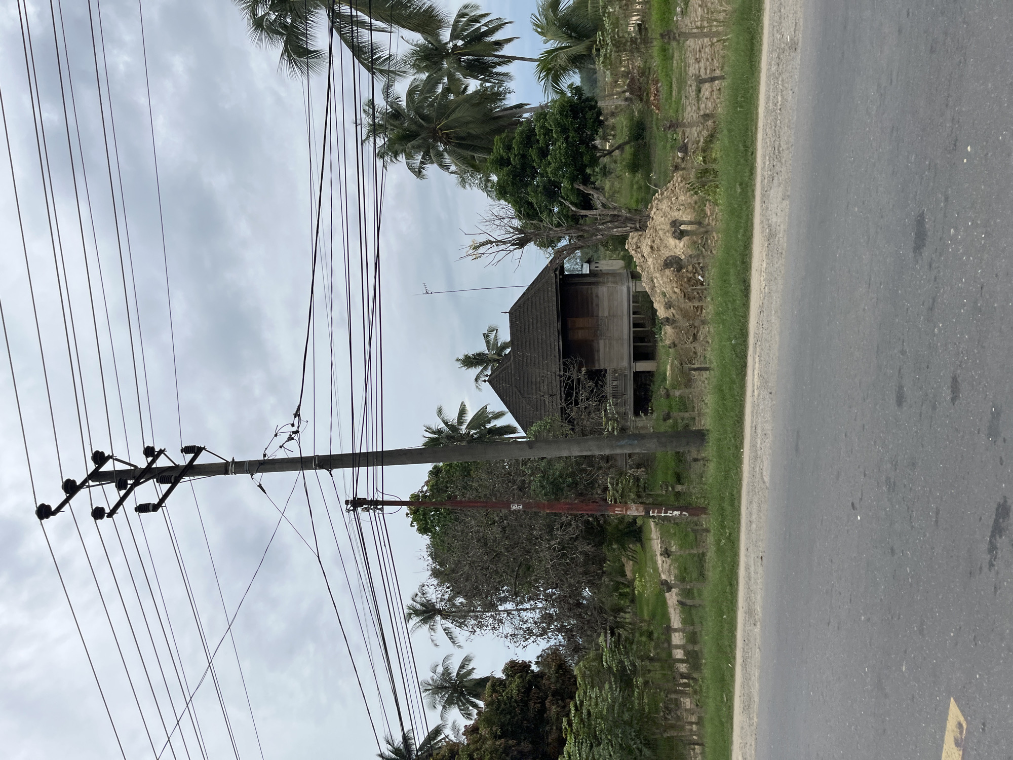 The image shows a rural scene in Simpangempat, Indonesia, with a paved road running along the right side. A power pole with numerous wires stretches across the left side of the frame. In the background, a wooden house with a dark roof is visible amongst lush greenery and tall palm trees. The sky is overcast, suggesting a cloudy day. The overall impression is one of a quiet, perhaps slightly weathered, tropical environment. There is no visible text in the image.