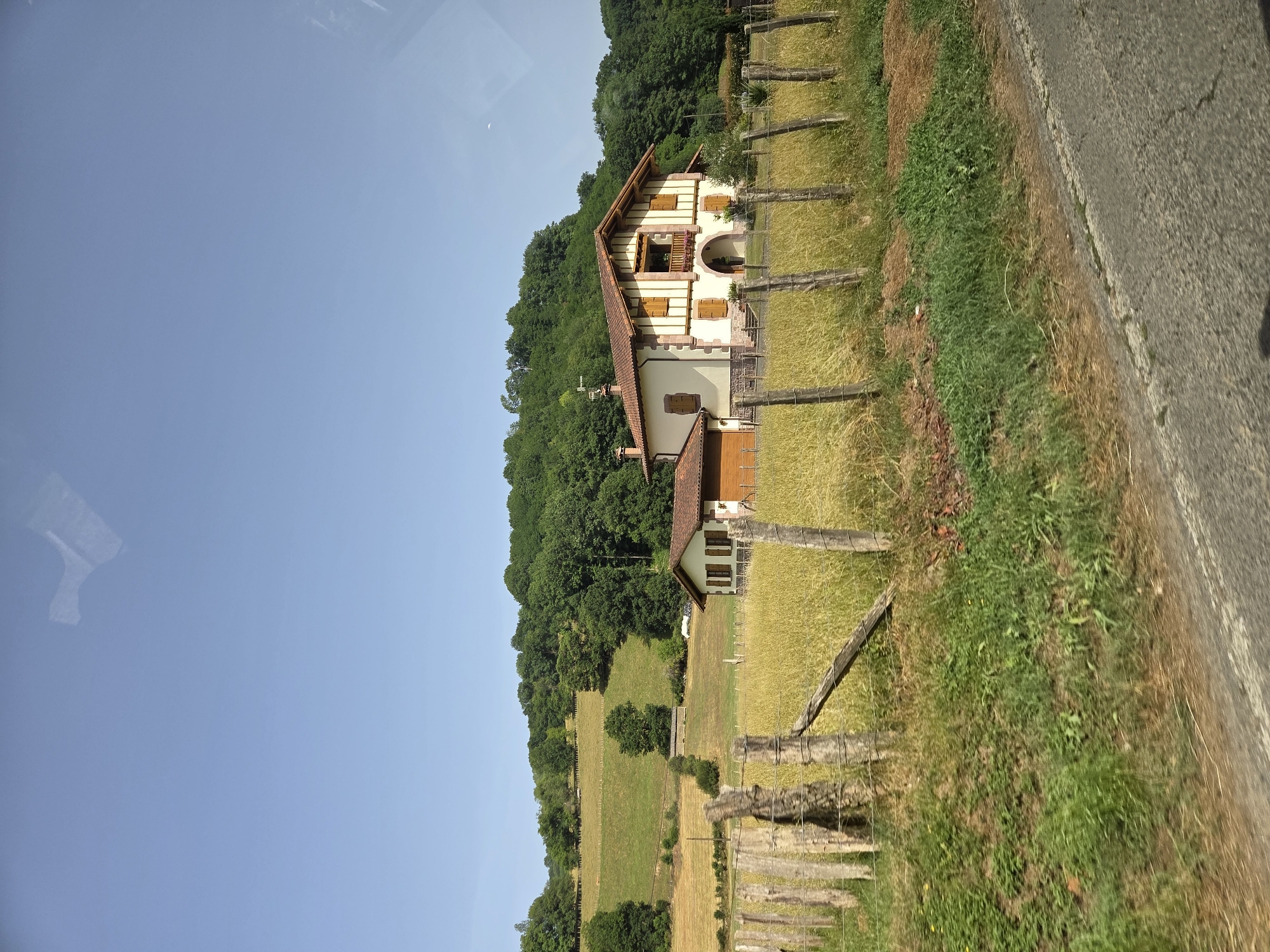 This image depicts a rural landscape in Baztan, Spain. The scene is dominated by a traditional white house with red-tiled roof, nestled on a hillside covered with lush green trees. The house features wooden shutters and a small balcony with floral decorations. In front of the house, a field of tall, golden grass stretches towards a rustic wooden fence. Beyond the field, rolling green hills are visible, also densely forested. The sky is a clear, bright blue, suggesting a sunny day. A faint crescent moon is visible in the sky. The image appears to have been taken from a moving vehicle, as indicated by the slightly blurred asphalt road on the right side of the frame and what looks like a reflection on the left side, possibly from a window. The overall impression is one of tranquility and natural beauty in a countryside setting. There is no visible text in the image.