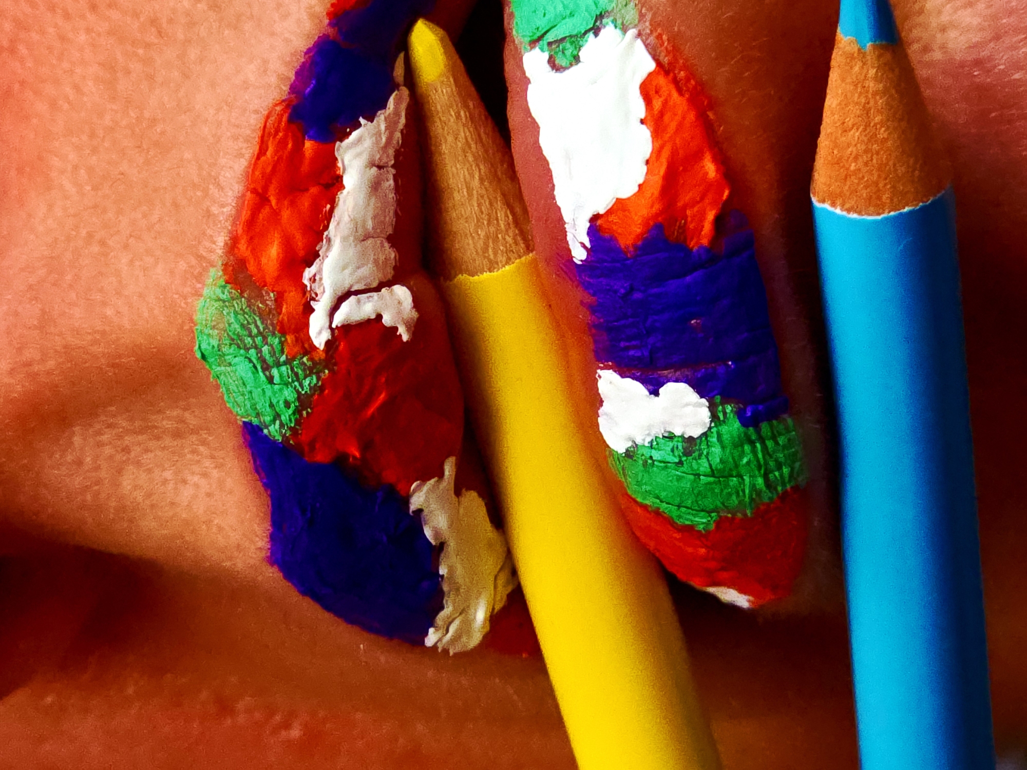 A close-up shows fingers covered in thick blotches of green, red, white, and blue paint, interspersed with a yellow and a blue colored pencil. The scene is set against a peach-toned, textured background.