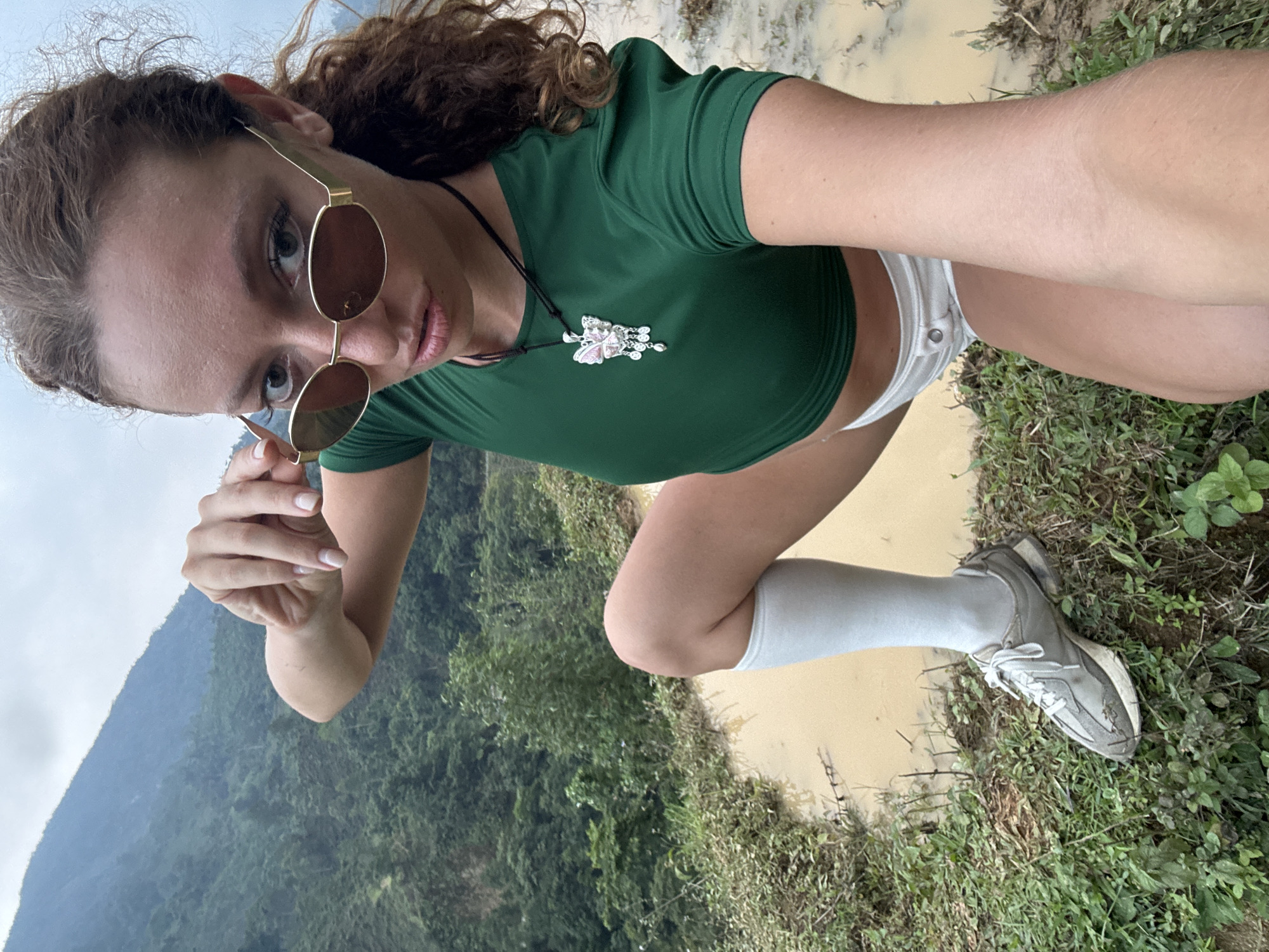 An eye-level selfie shows a young woman posing in a natural, mountainous setting in Nậm Dịch Commune, Vietnam. She is crouched down, leaning slightly to her left, looking directly at the camera. She has long, curly reddish-brown hair pulled back, and is wearing round, gold-rimmed sunglasses, which she holds slightly down on her nose with her left hand, revealing her eyes.
She is dressed in a dark green, short-sleeved crop top that exposes her midriff, white shorts, white knee-high socks, and light-colored sneakers, which appear somewhat worn or muddy. A distinctive silver-toned pendant necklace, possibly depicting a butterfly or floral design with pink and white elements, hangs from a black cord around her neck. Her lips are slightly parted.
The background features lush green foliage and trees, with hazy mountains under an overcast sky. To her right, there is a muddy or watery area, possibly a puddle or a small stream, next to a grassy patch. The overall impression is one of an outdoor adventure or trek on a cloudy day. No visible text is present in the image.